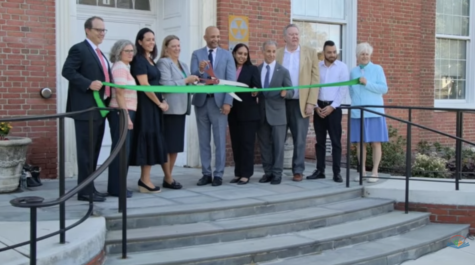 A group of ten people stand on the steps of a red brick building for a ribbon-cutting ceremony, with one man using large scissors to cut a green ribbon.