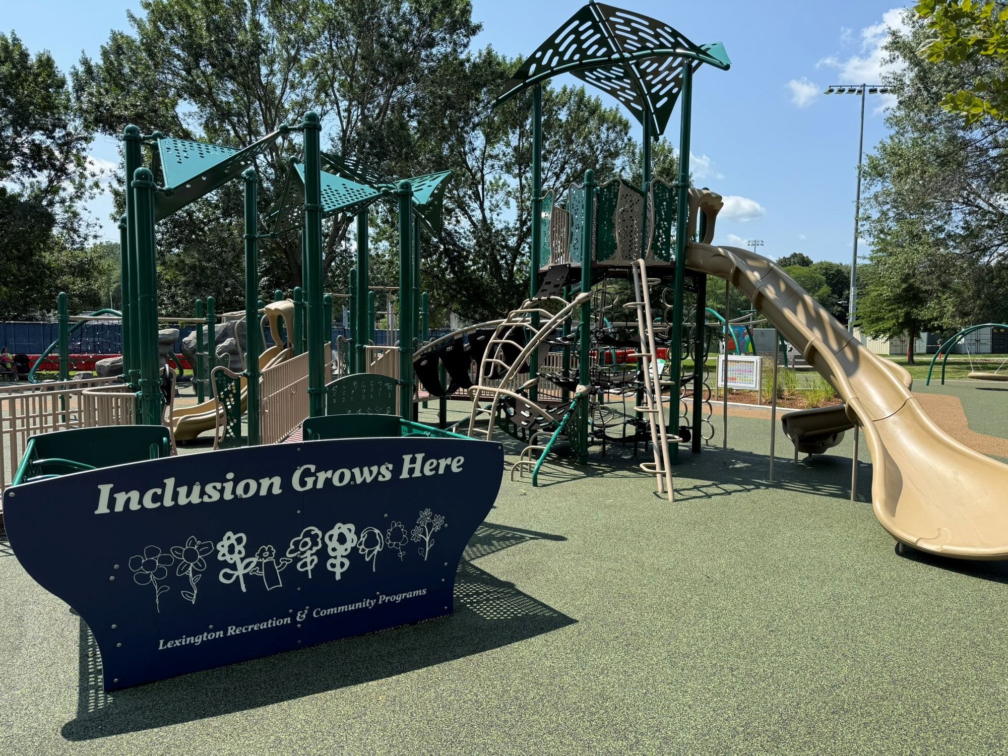 Green playground climbing structures with a sign reading 'Inclusion Grows Here' on rubber safety surfacing.