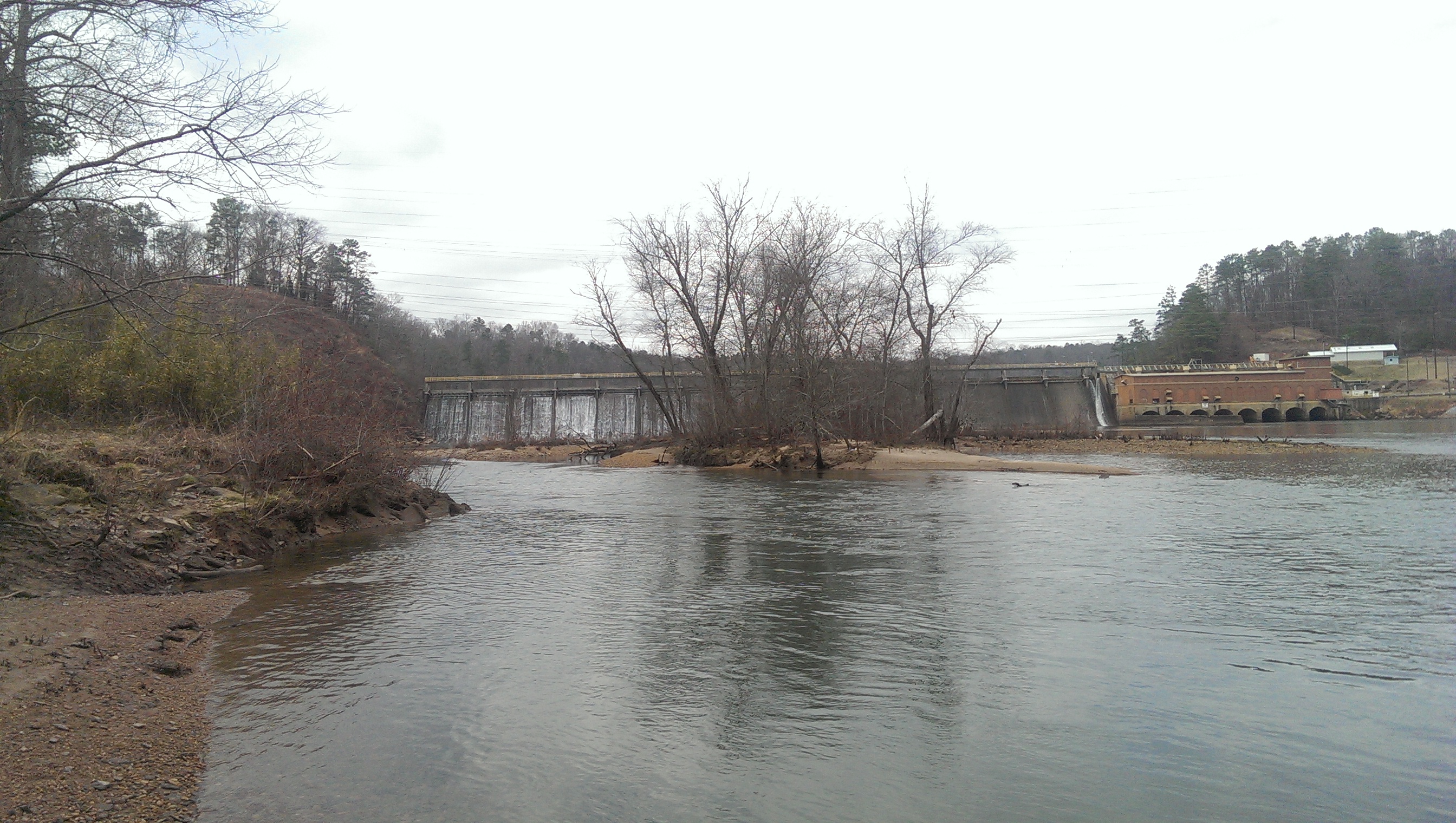 Concrete dam spanning the Chattahoochee River with bare trees along a sandy bank.