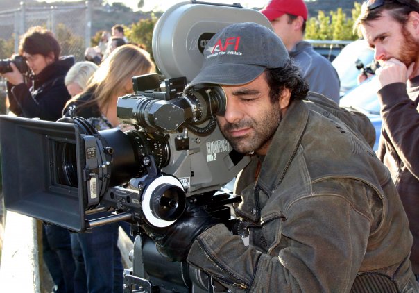 A person in a cap peers through a professional cinema camera on an outdoor film set.