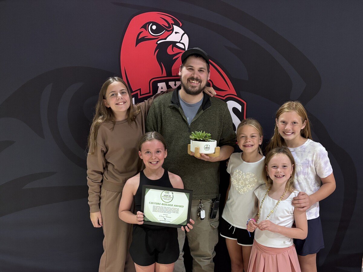 A man holding a potted plant poses with five smiling children in front of a school mascot mural.