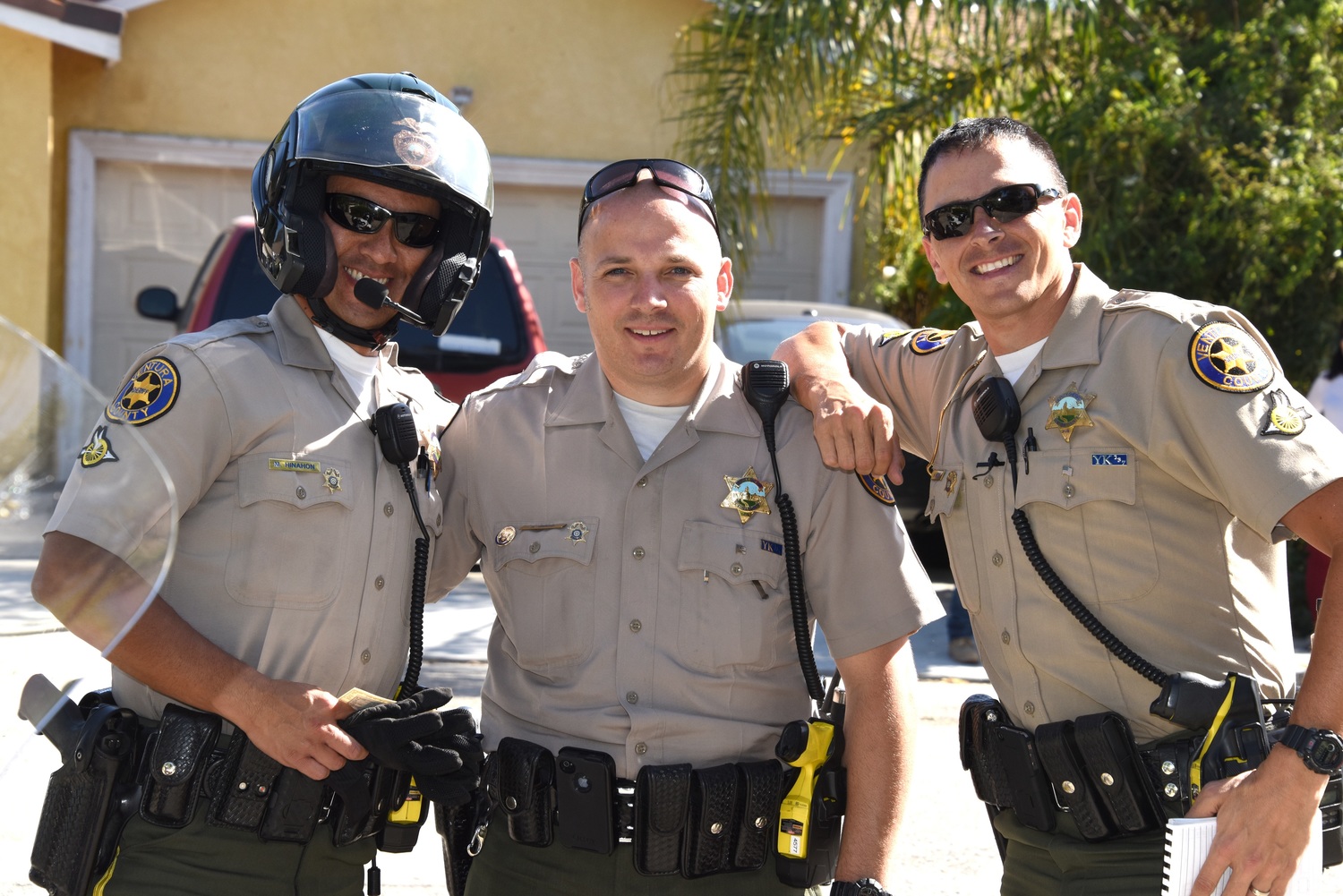 Three uniformed sheriff's deputies pose together outdoors, one wearing a motorcycle helmet.