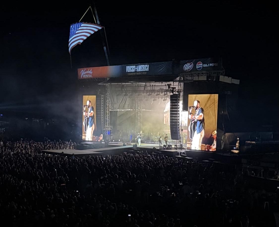 Outdoor concert stage at night with large LED screens and a massive crowd.