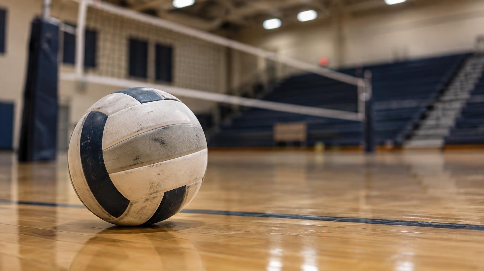 A volleyball resting on a hardwood gym floor near a net, bleachers blurred behind.