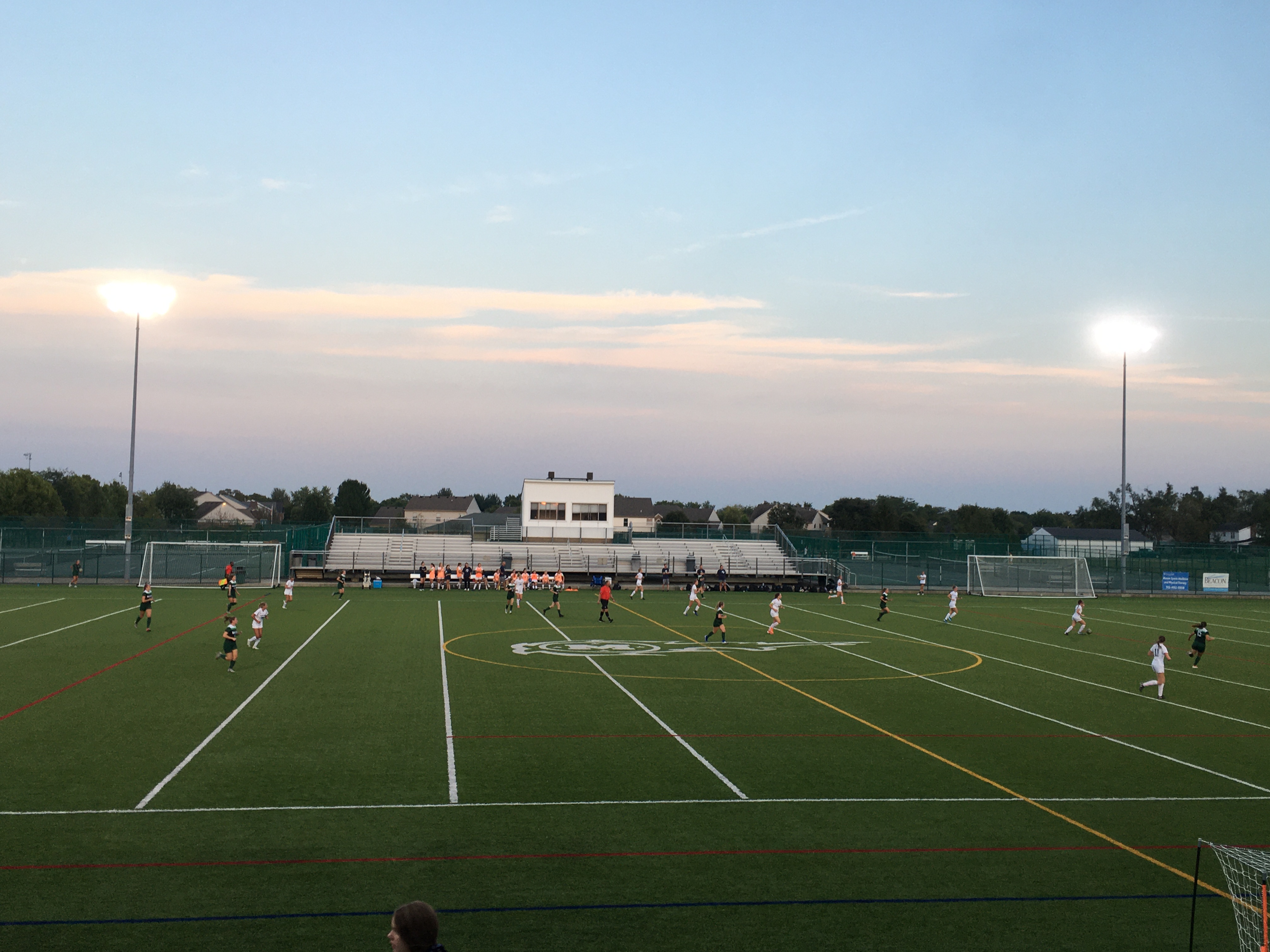 Floodlit artificial turf stadium at dusk with bleachers and a press box in the background.