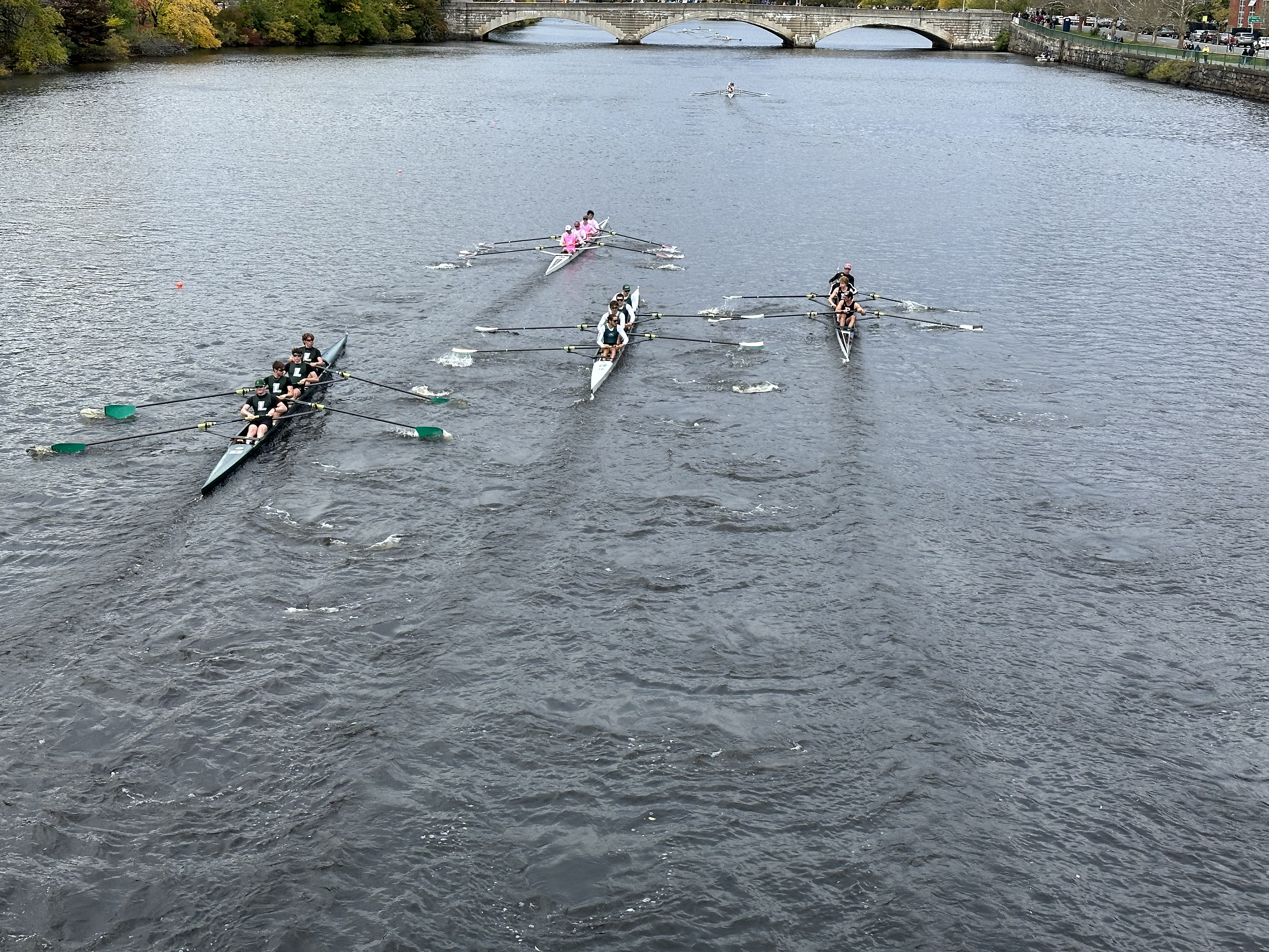 Four rowing shells competing upstream on the Charles River, viewed from a bridge above.