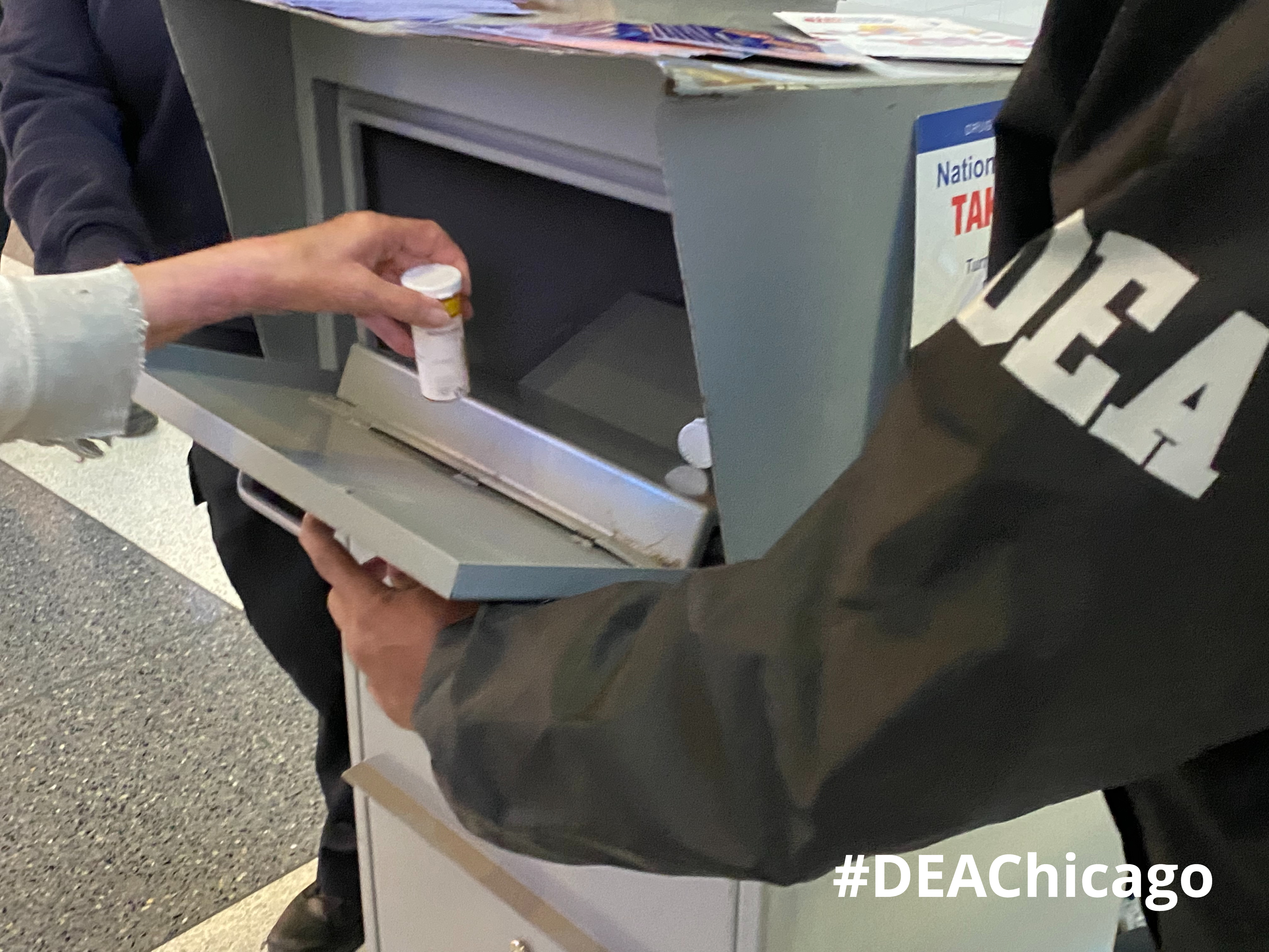 A person deposits a prescription pill bottle into a gray drug collection box.