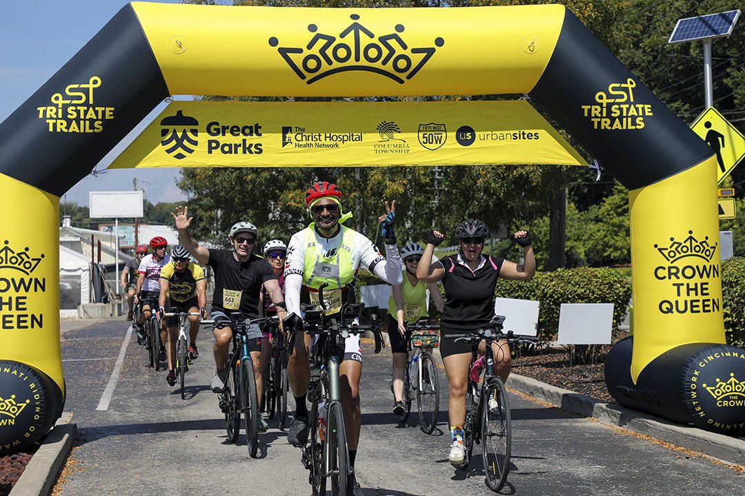 Cyclists raise their arms in celebration while crossing under a large yellow inflatable event arch.