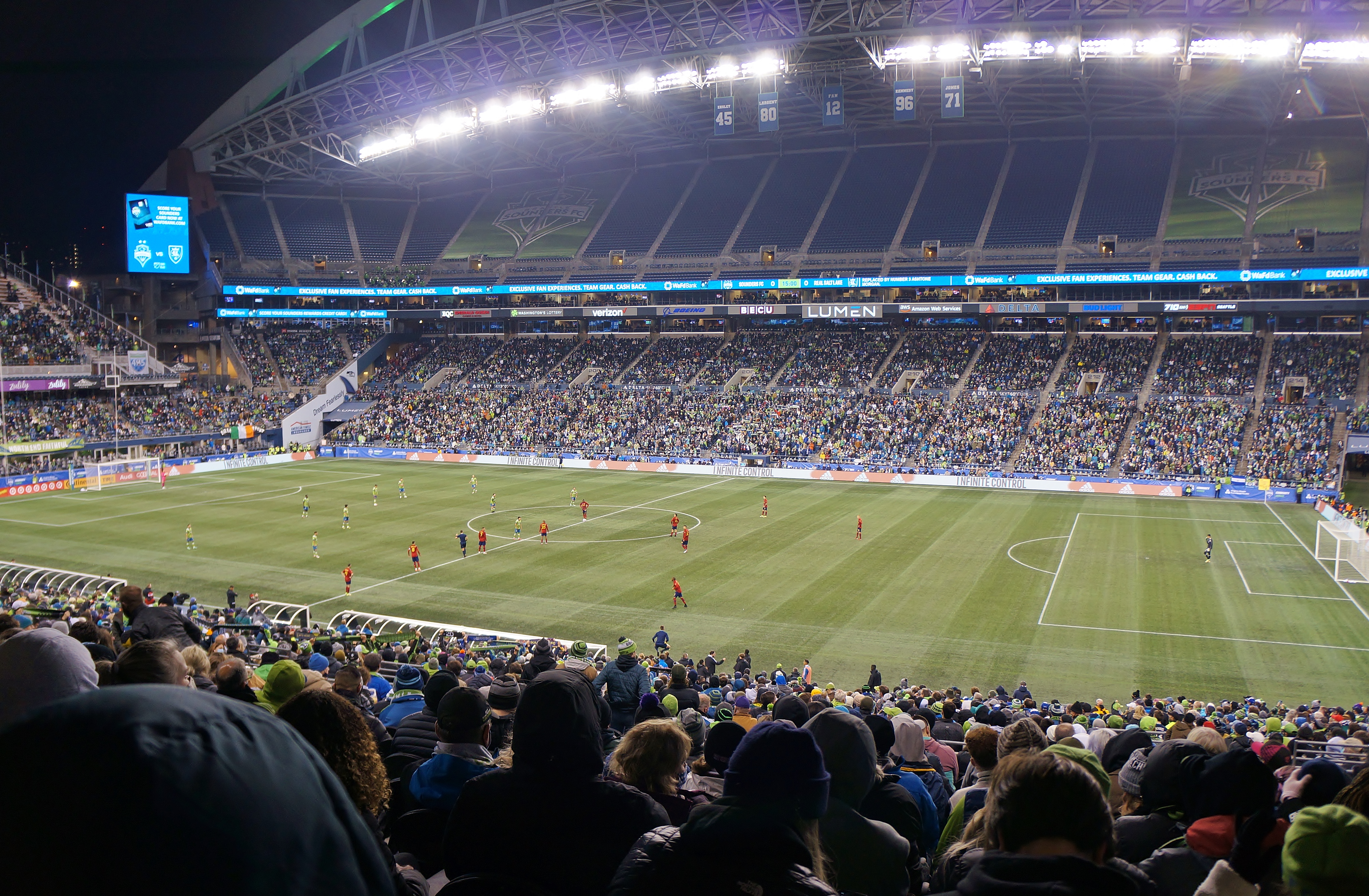Soccer players on a green pitch inside a large, packed stadium at night.