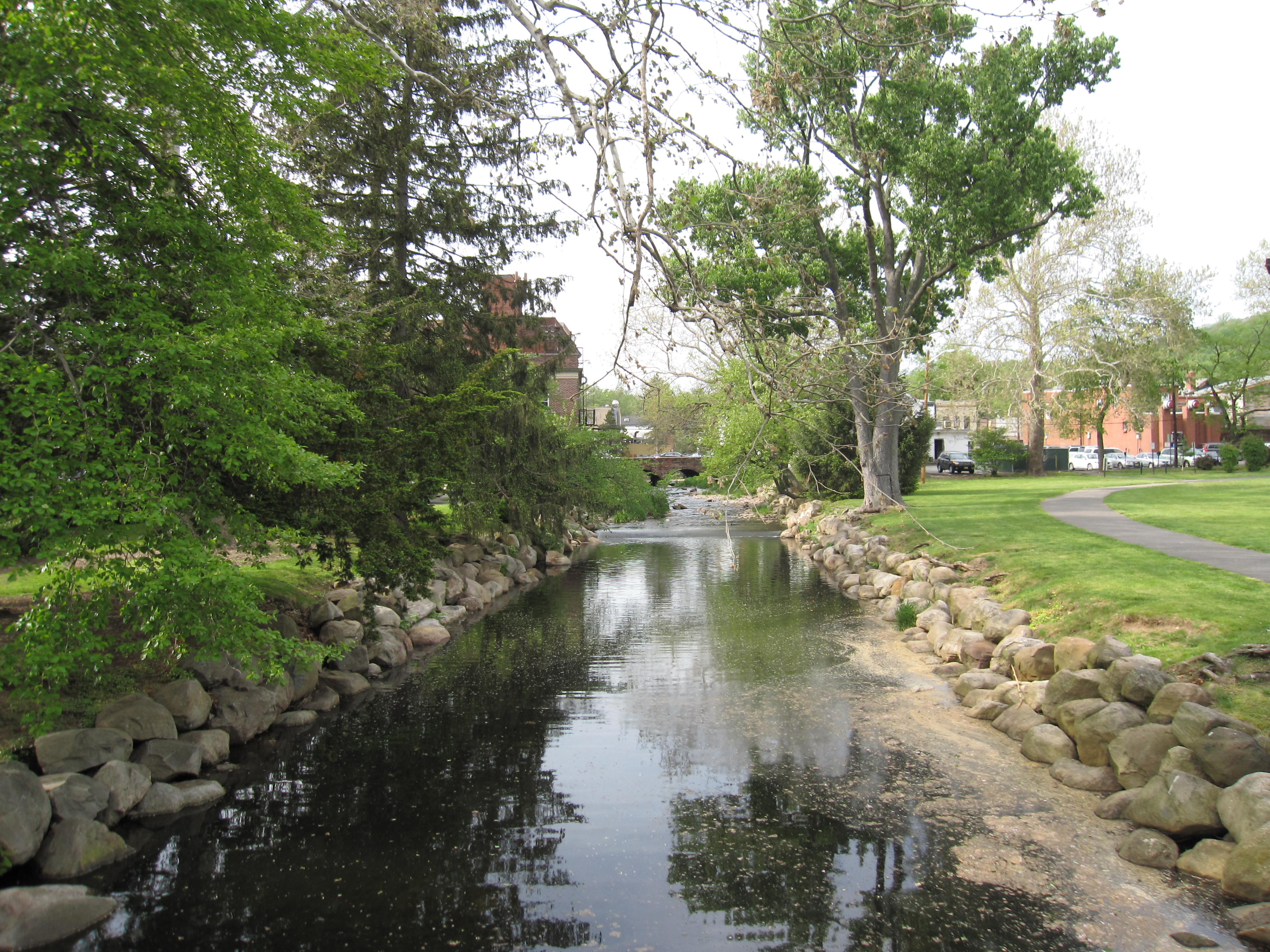 A narrow brook with stone-lined banks flows through a tree-lined park.