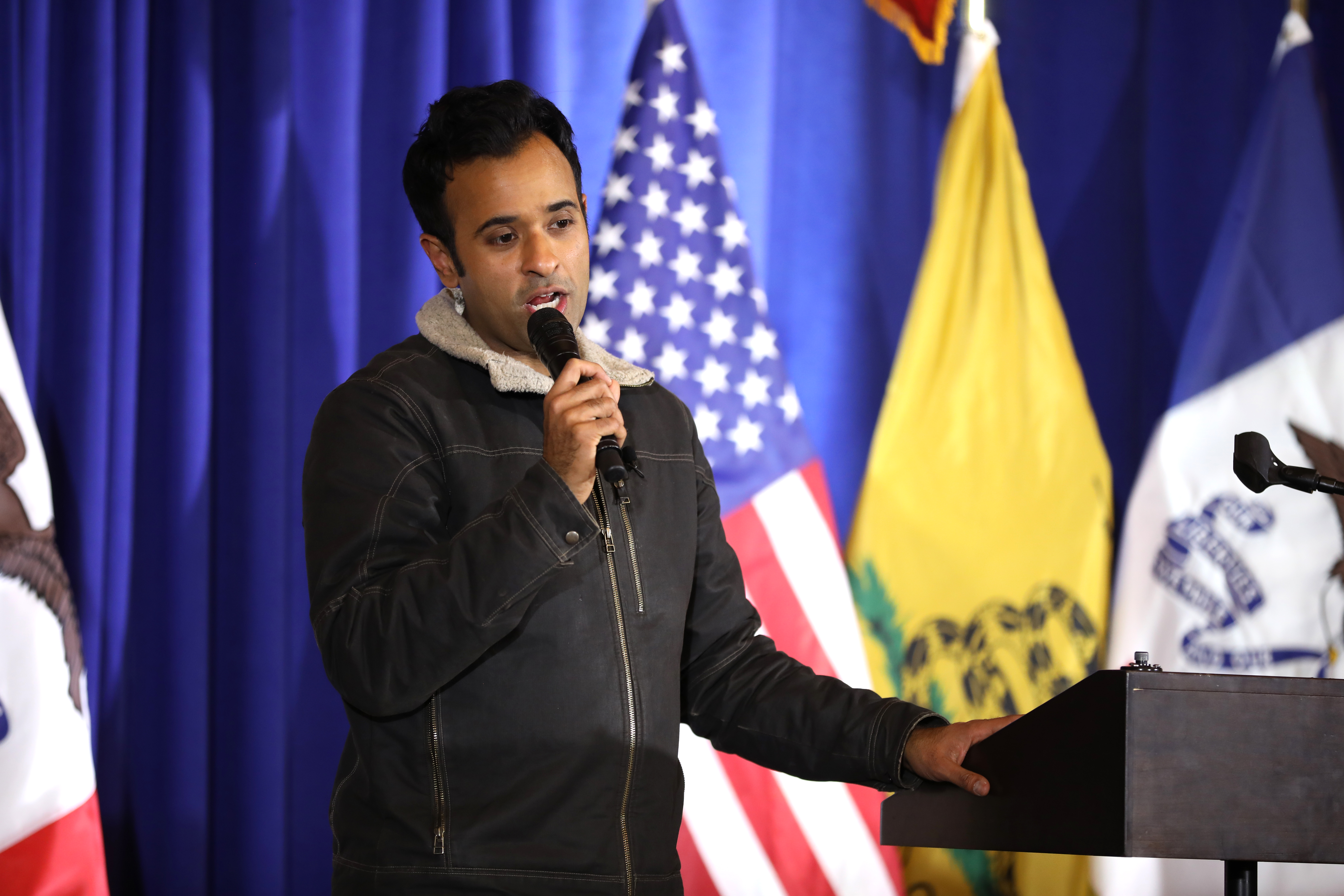 Man speaking into a handheld microphone at a podium with American flags behind him.