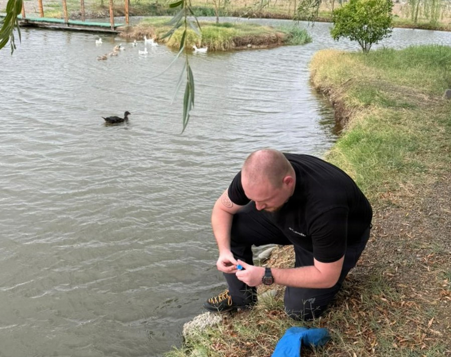 Person crouching at a pond's edge collecting a water sample in a small vial.