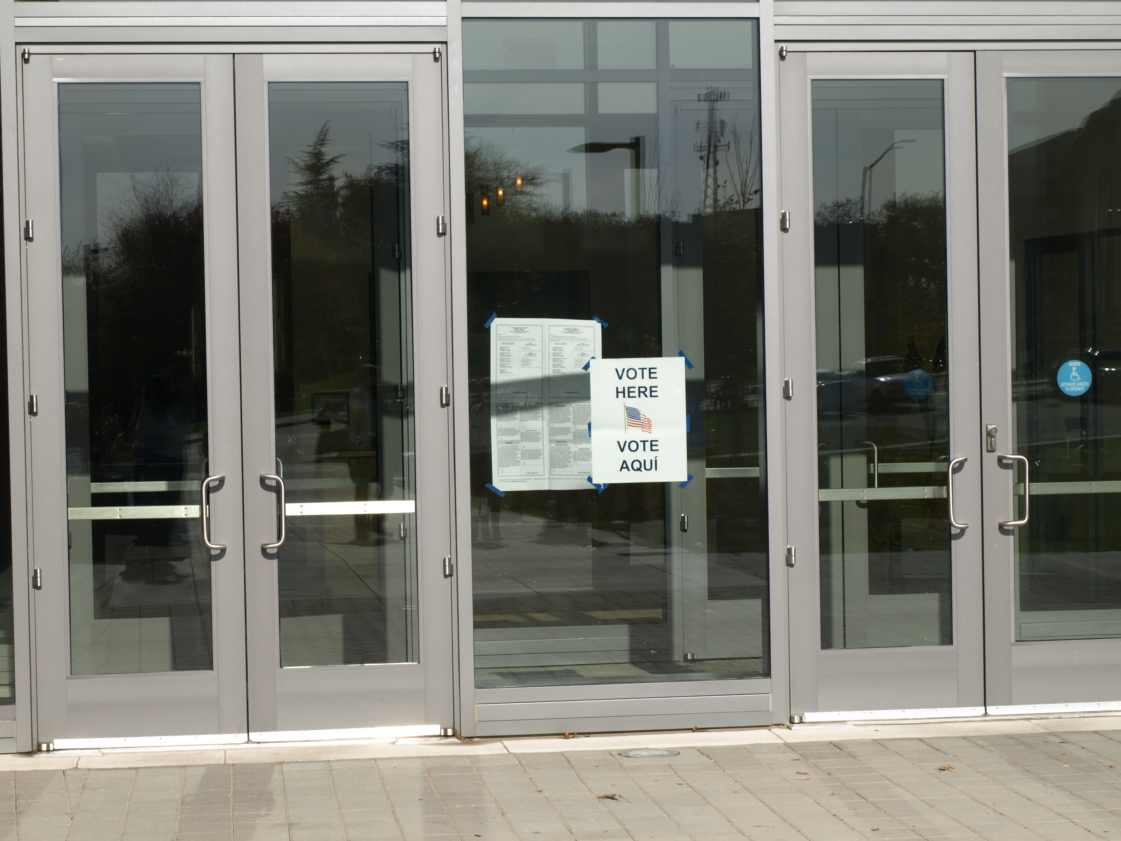 Glass building entrance with a bilingual Vote Here and Vote Aqui sign posted on the door.