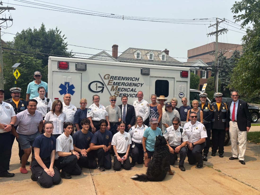 Large group of EMS personnel and civilians posing in front of an ambulance outdoors.
