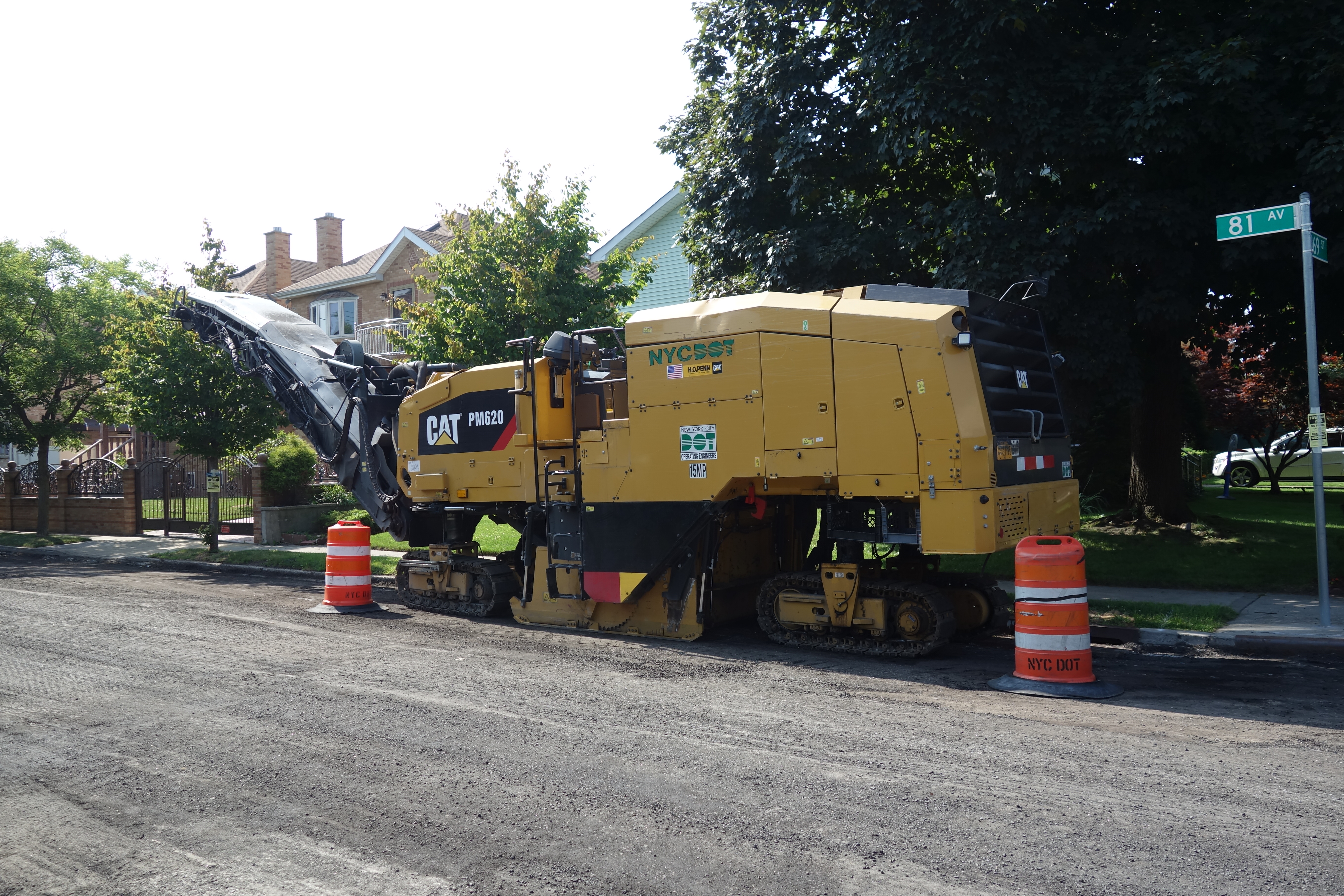 Yellow Caterpillar cold planer machine milling a residential street with orange traffic barrels.