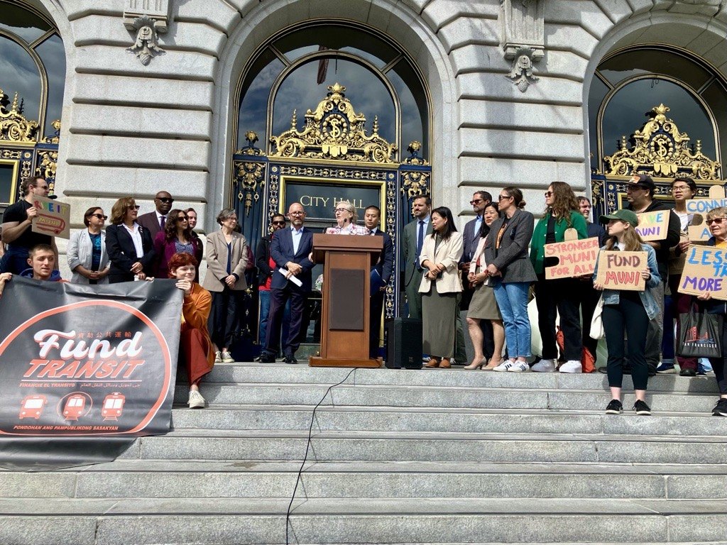 Crowd gathered on steps of a city hall building, some holding transit-advocacy signs, with a speaker at a podium.