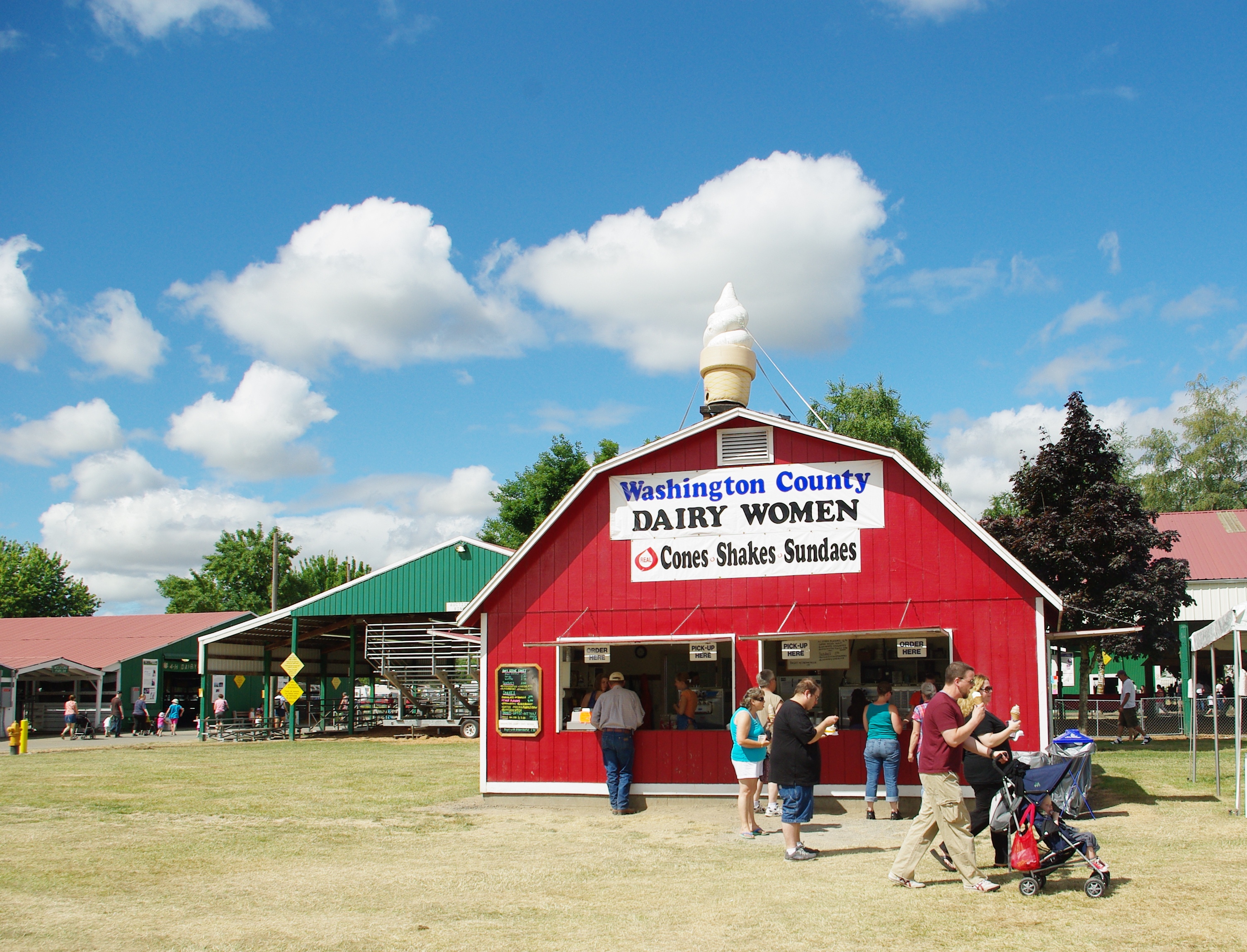 Red barn-style building with a large ice cream cone on the roof and fairgoers gathered in front on a sunny day.