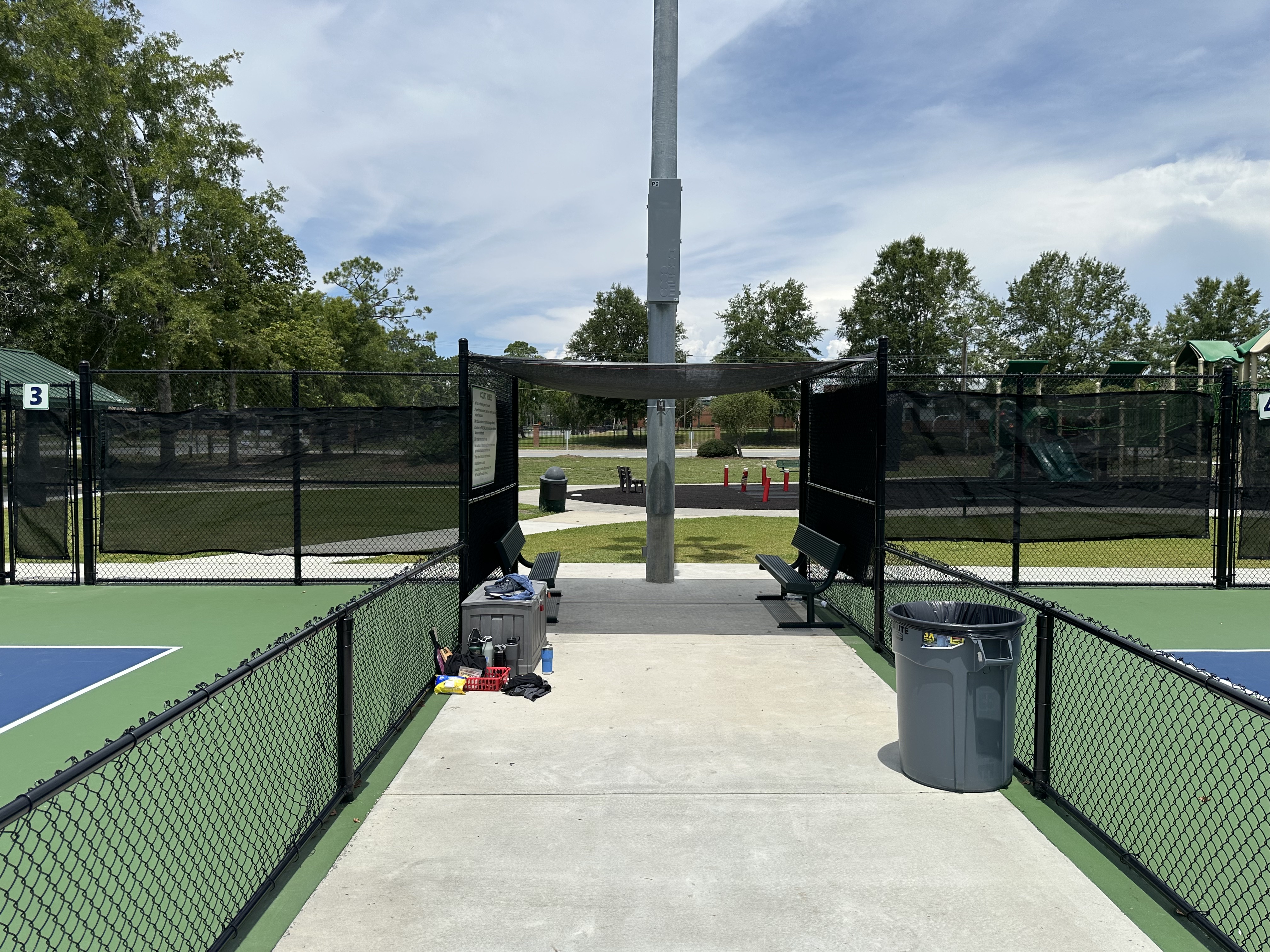 Walkway between two fenced outdoor pickleball courts with benches and a storage bin.