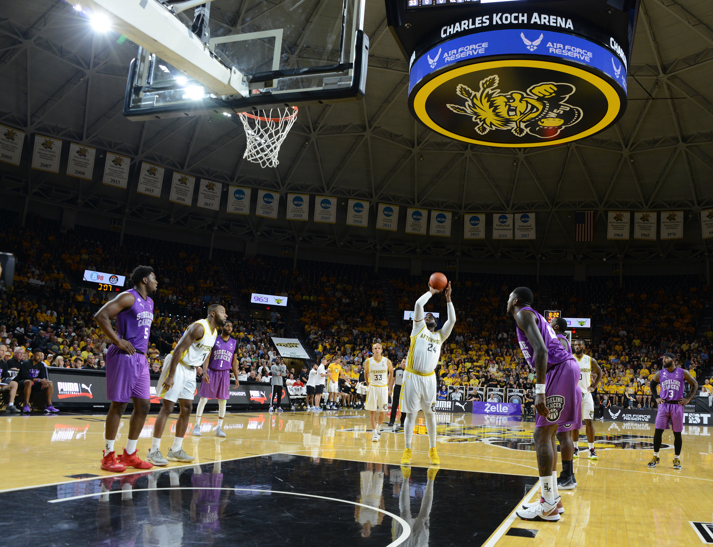 Packed indoor basketball arena with players on the court and a large overhead scoreboard.