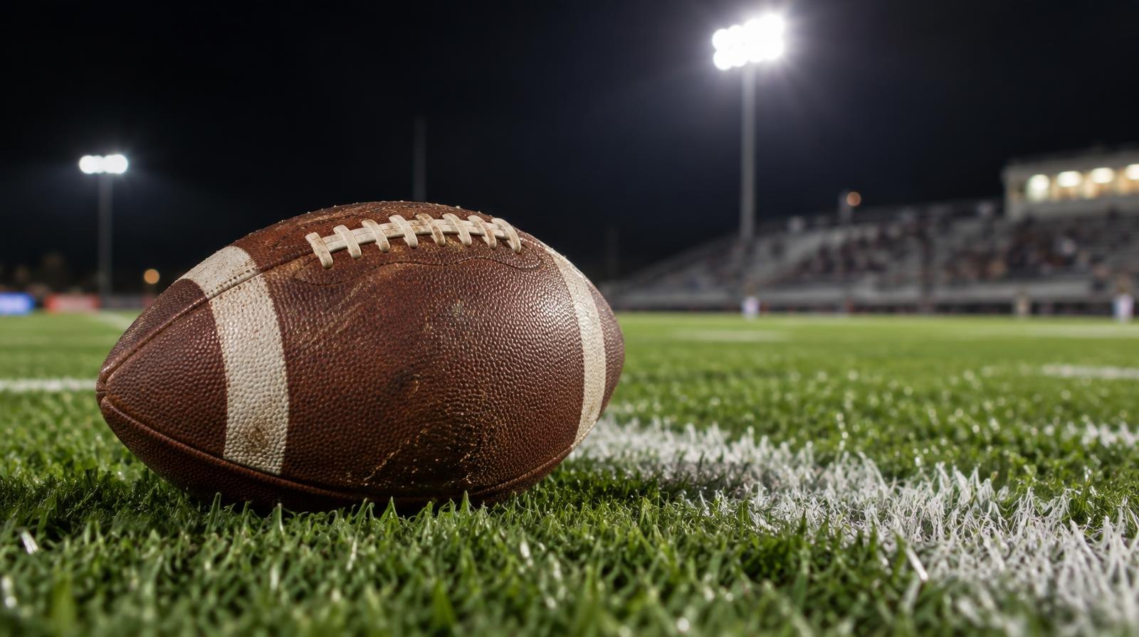 An American football sitting on green grass near a white yard line under bright stadium lights.
