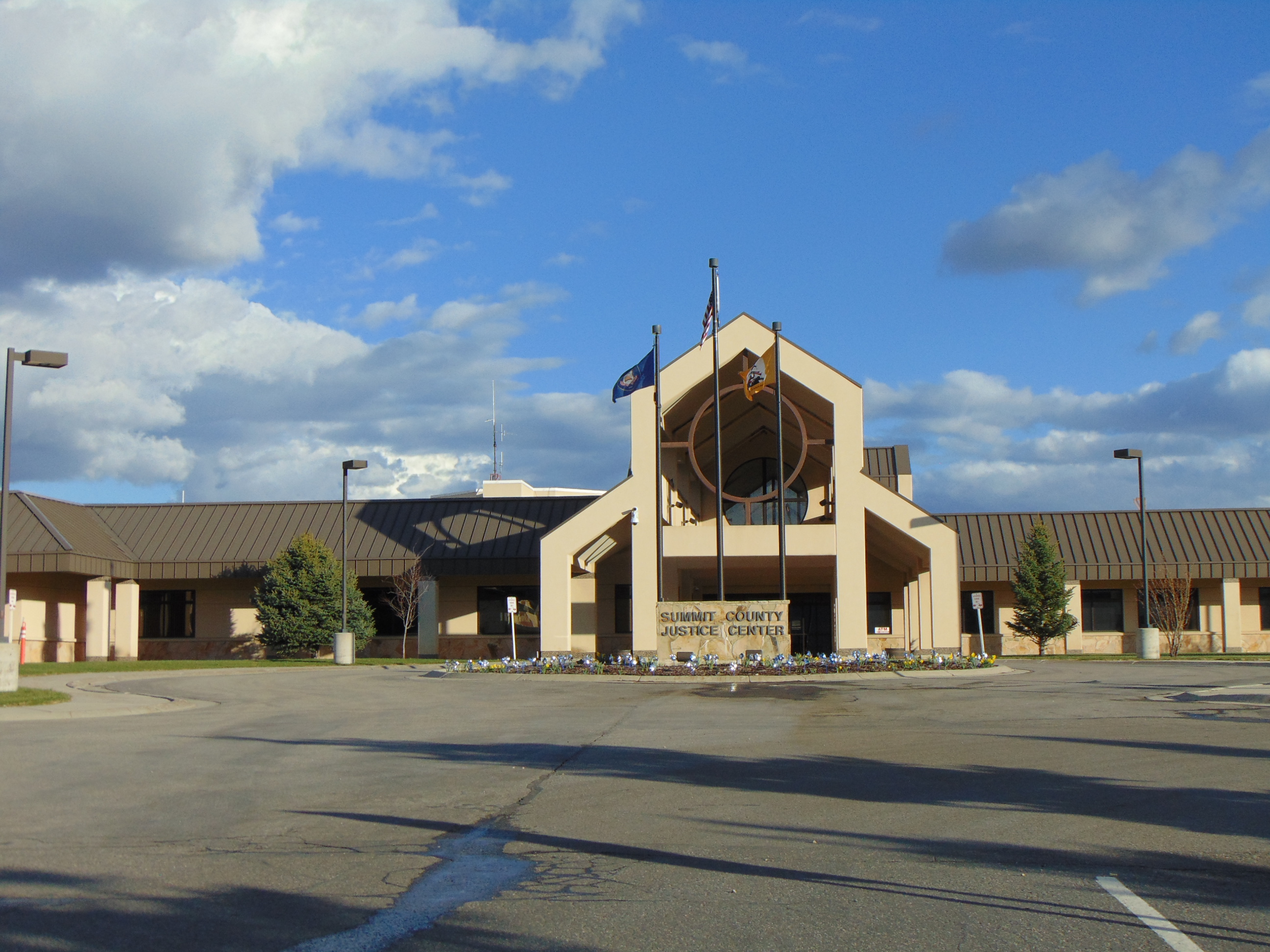 Tan building with peaked entrance and two flagpoles in a large parking lot under a partly cloudy sky.