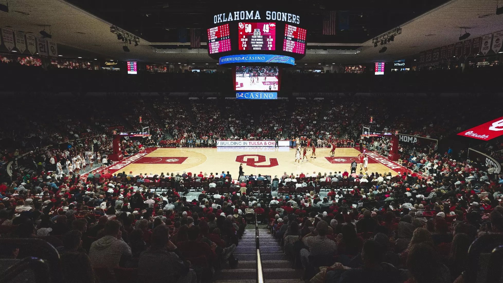 Wide interior view of a packed basketball arena with red-clad fans and players on the court.