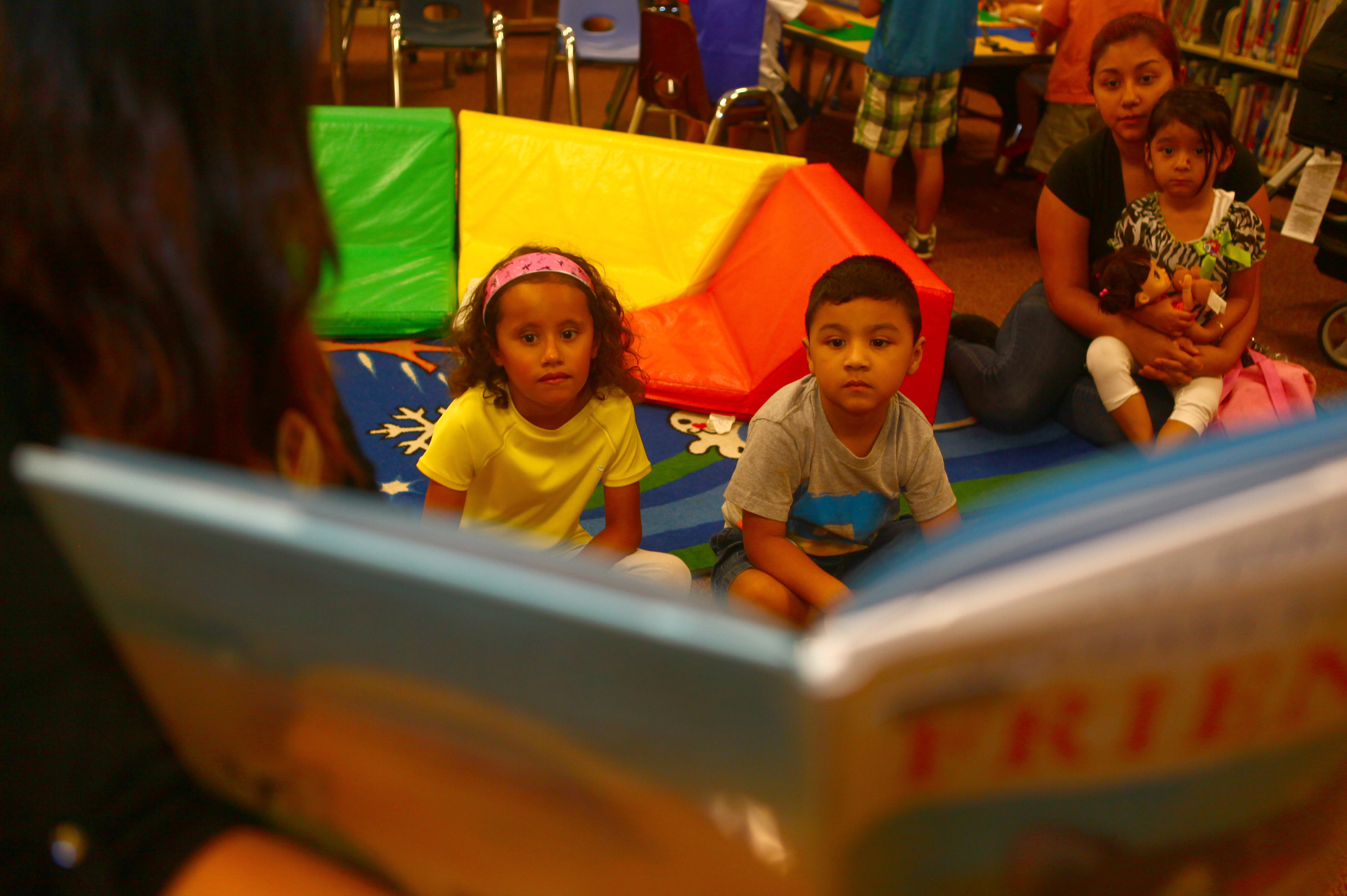 Young children sit on a library floor listening to a story being read aloud.