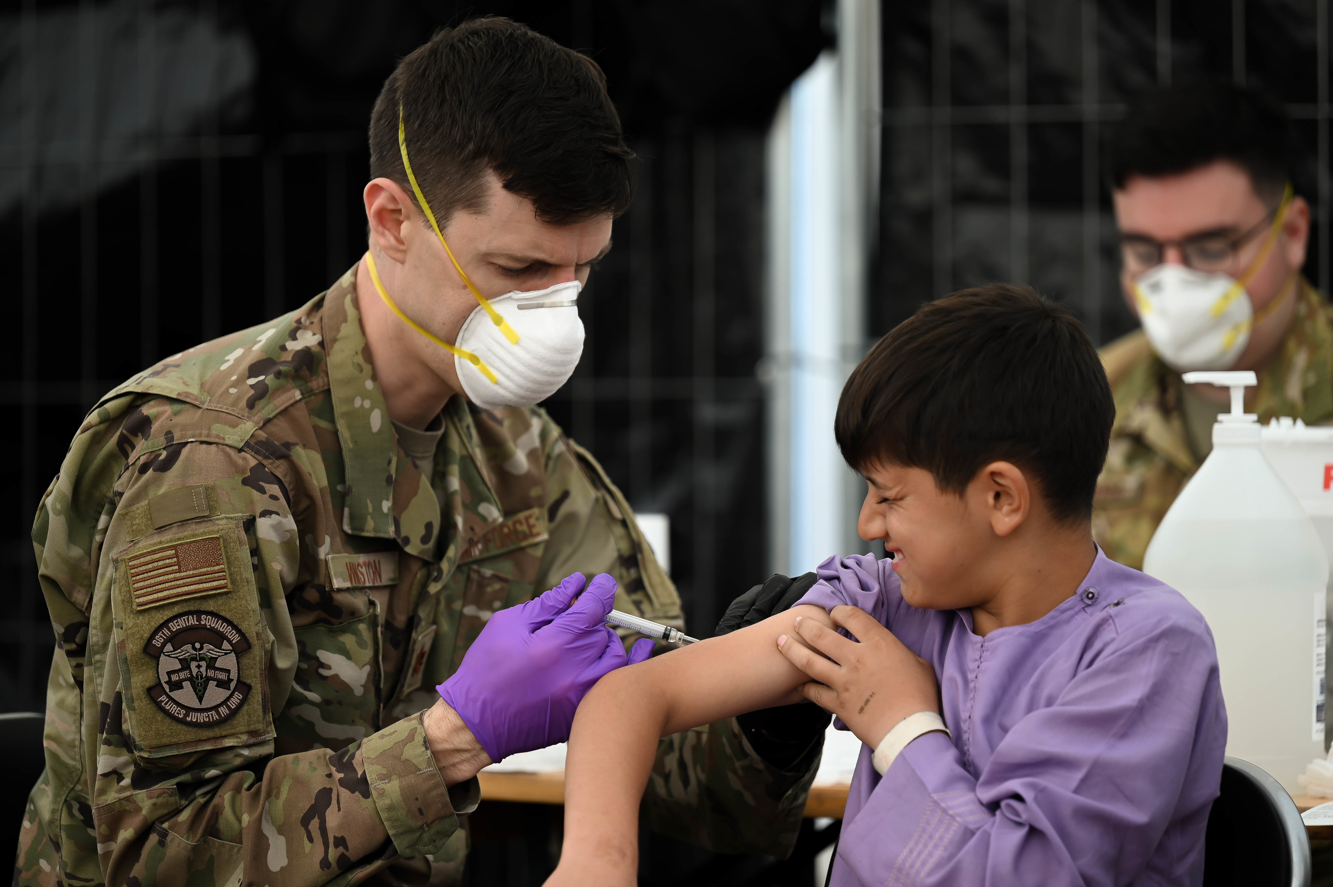 A masked medical worker in uniform gives a vaccine injection to a child's arm.