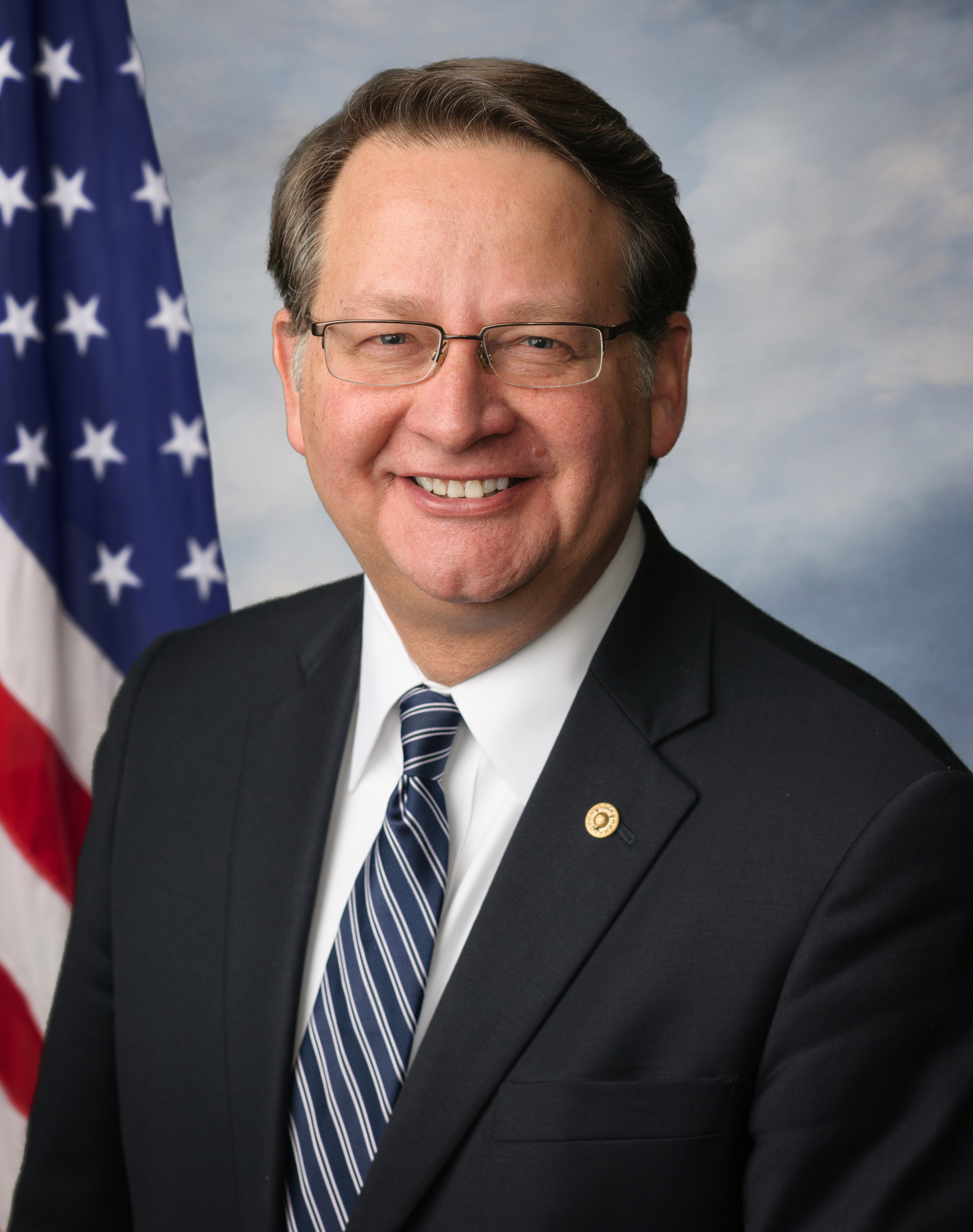 Man in dark suit and glasses smiling before an American flag backdrop.