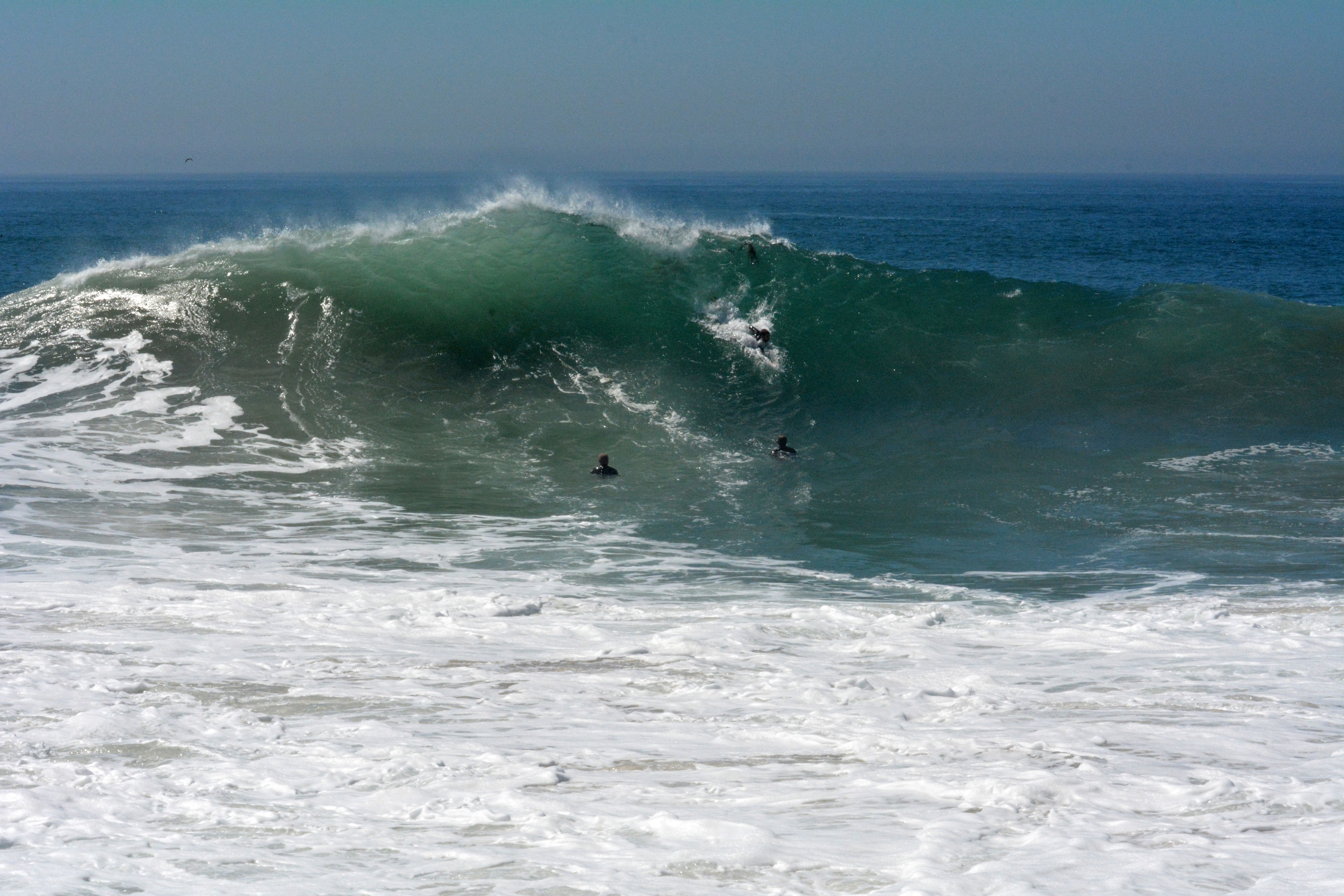 A large breaking wave crashes near shore at a sandy beach.