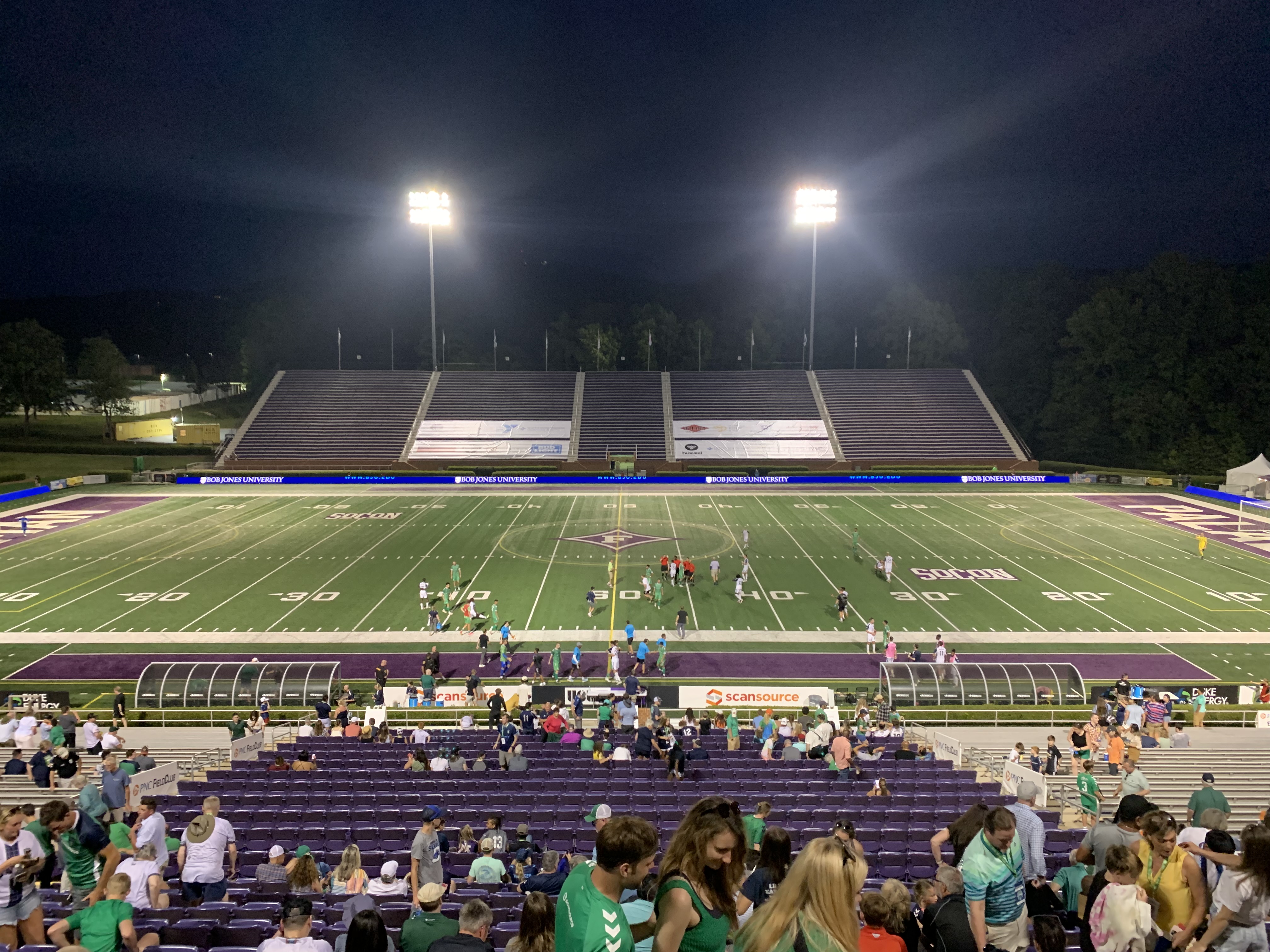 Floodlit soccer stadium at night with green field, purple seats, and spectators in the stands.
