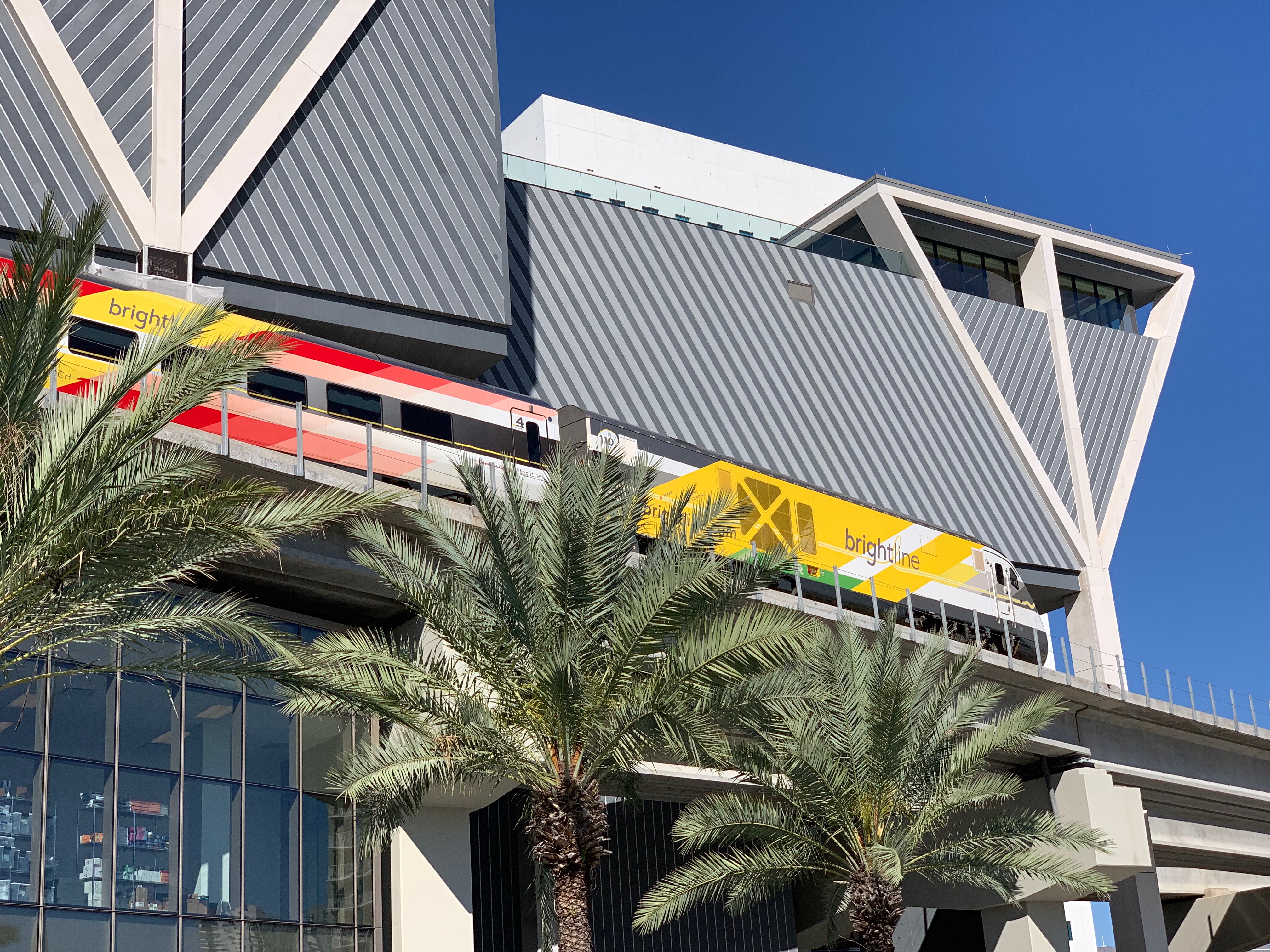 Brightline train alongside a modern angular station building with palm trees.