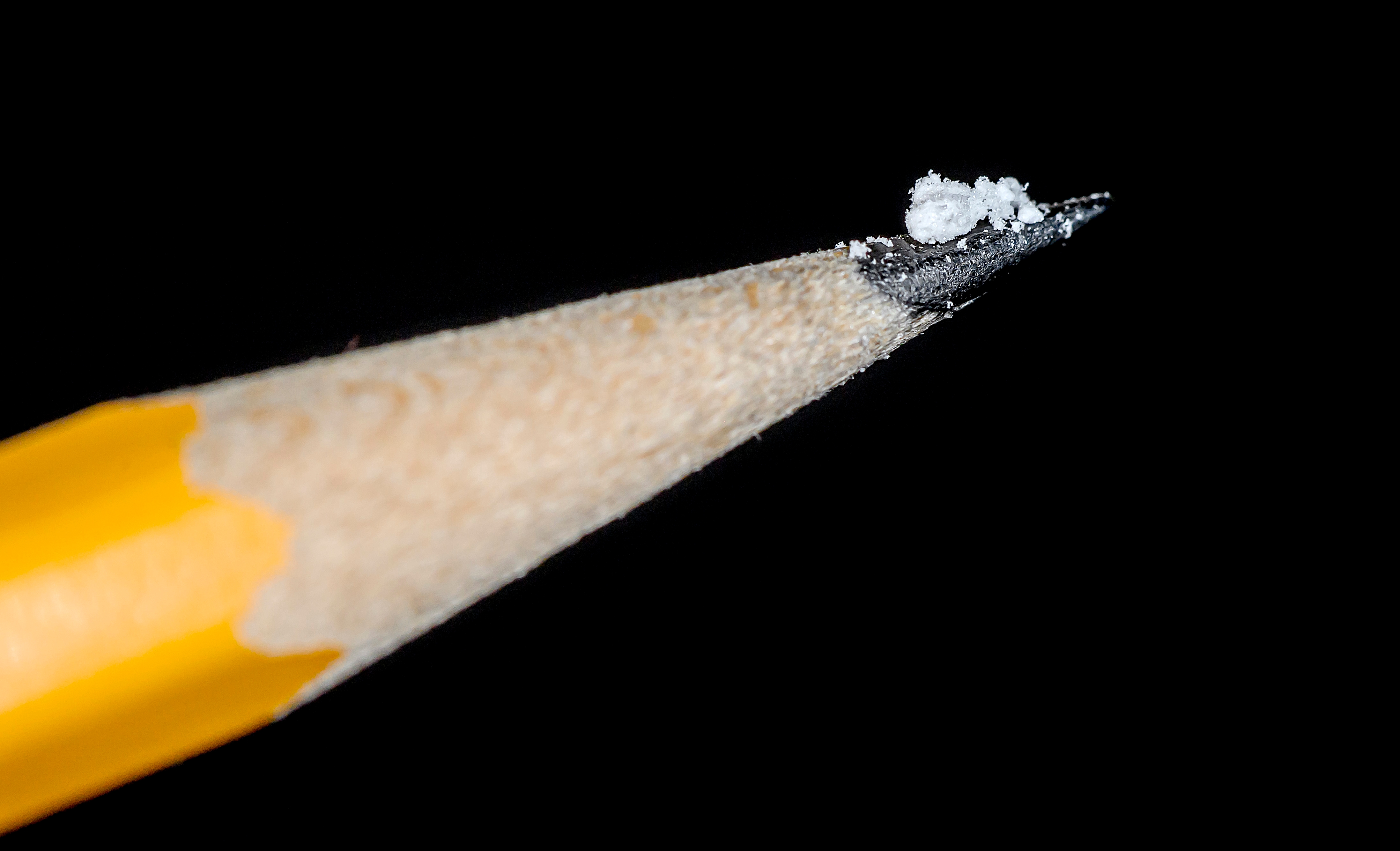 Macro photo of a tiny white crystalline substance on a yellow pencil tip against a black background.