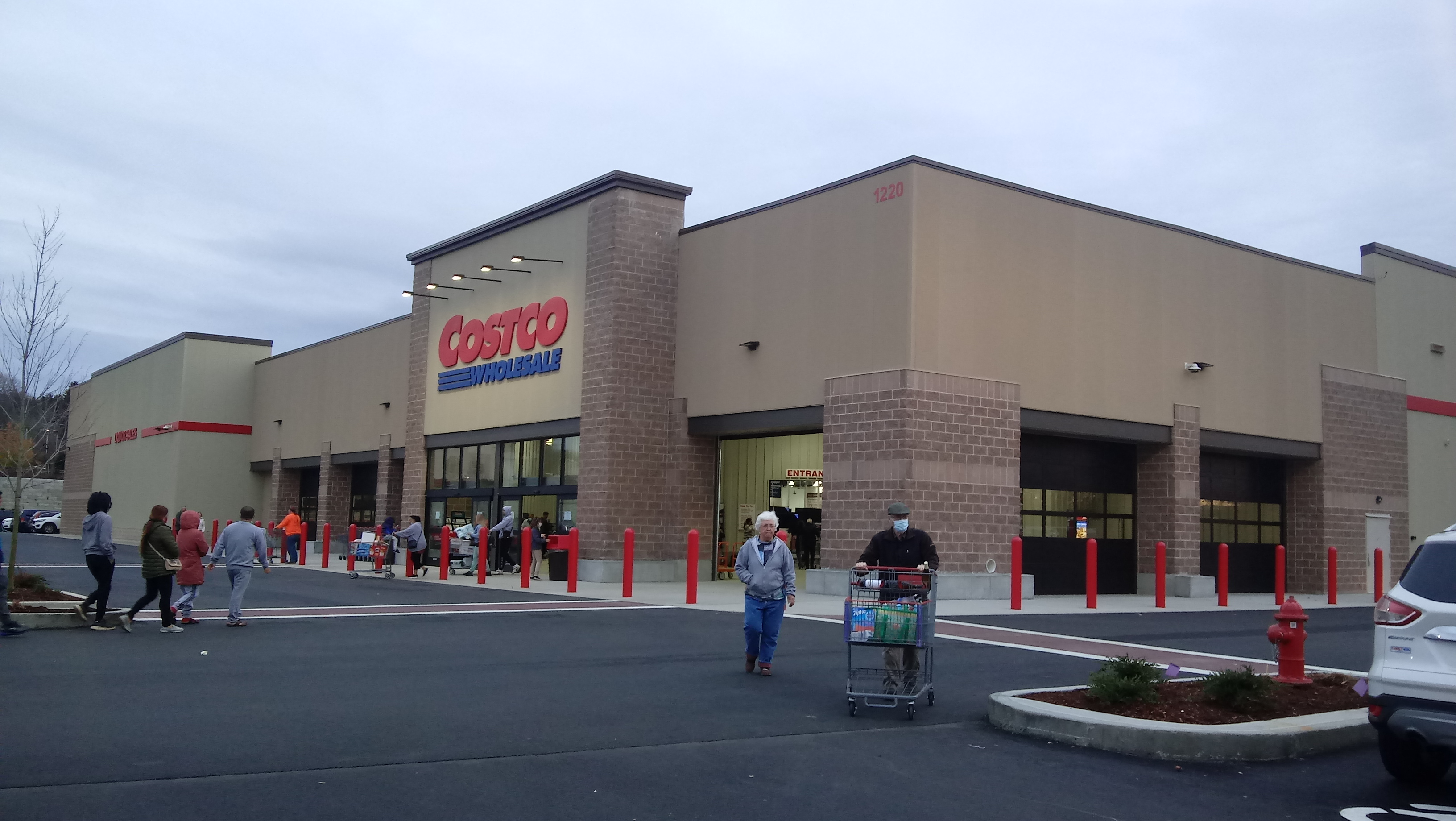 Exterior of a Costco Wholesale warehouse store with customers and shopping carts in the parking lot.