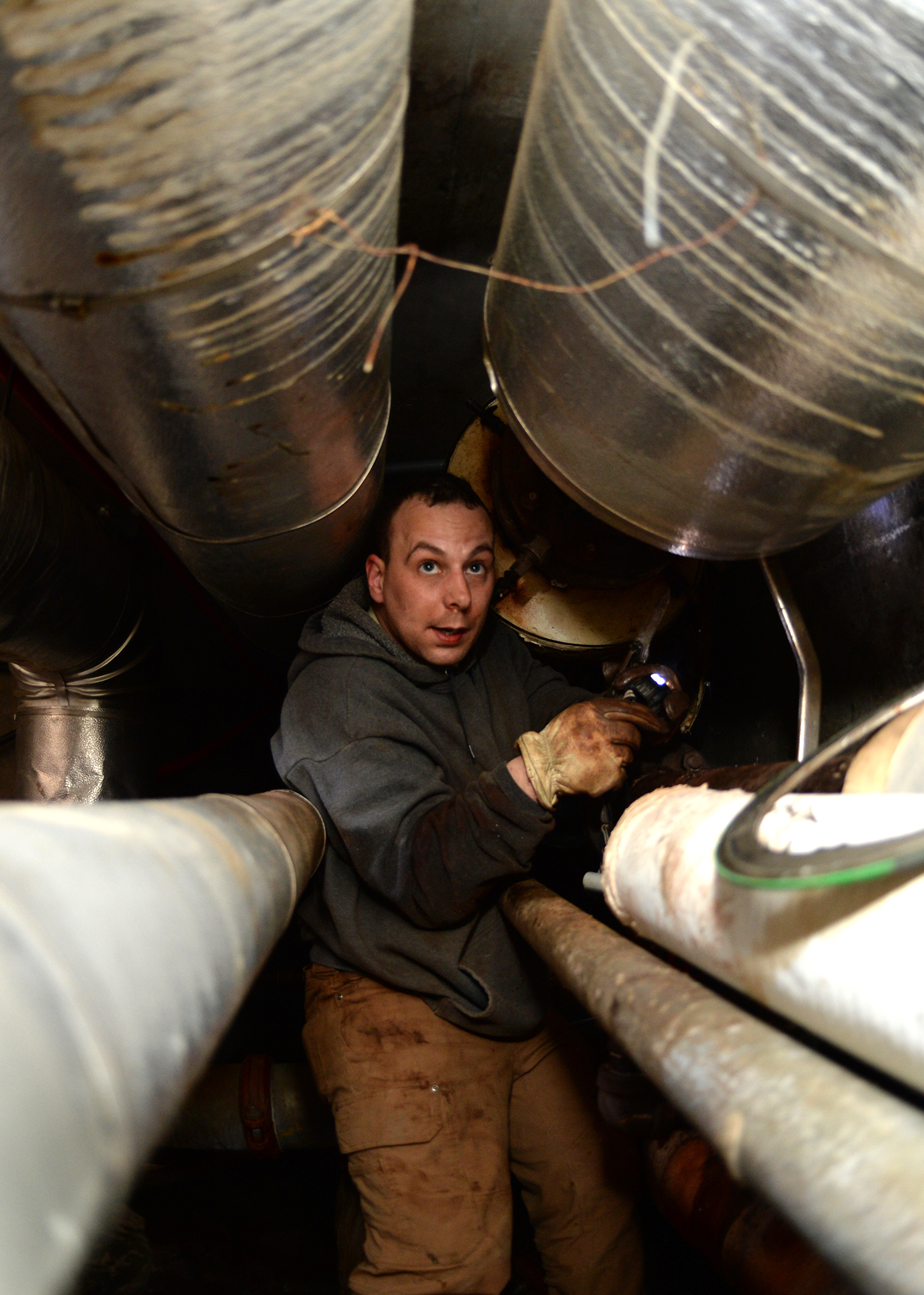 Worker crouched between large pipes in a narrow underground utility corridor.