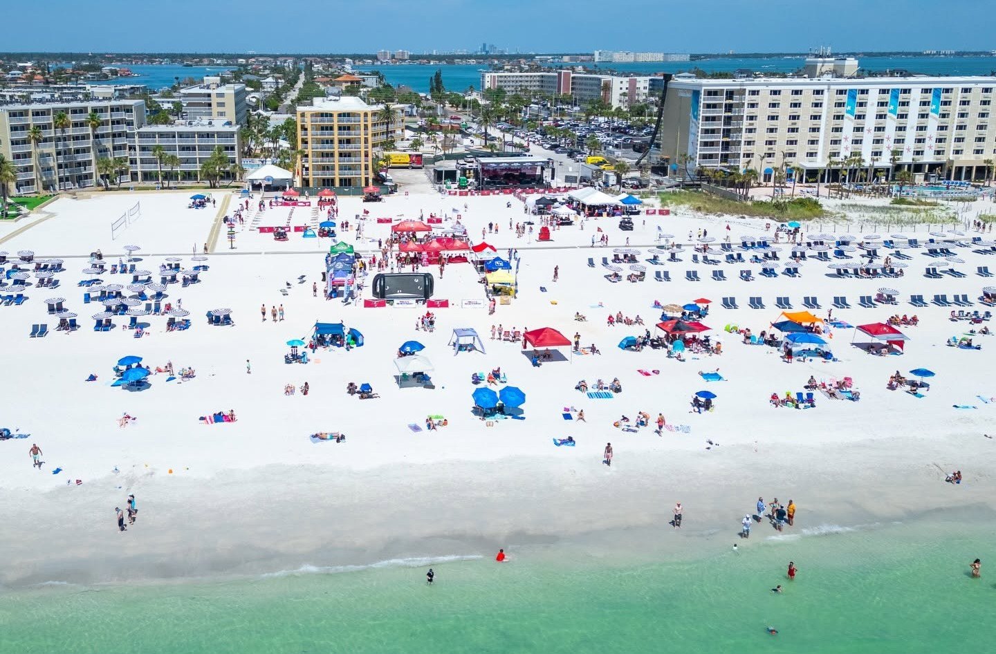 Aerial view of a crowded white-sand beach with colorful umbrellas, tents, and hotel buildings.