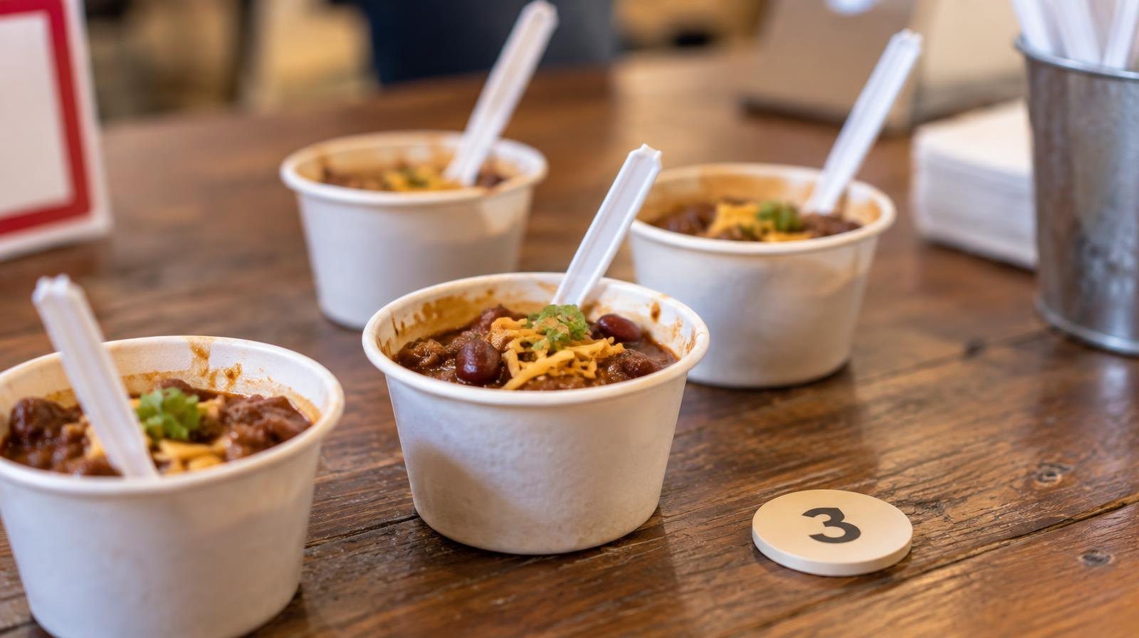 Small white cups of chili with spoons arranged on a wooden table beside a numbered chip.