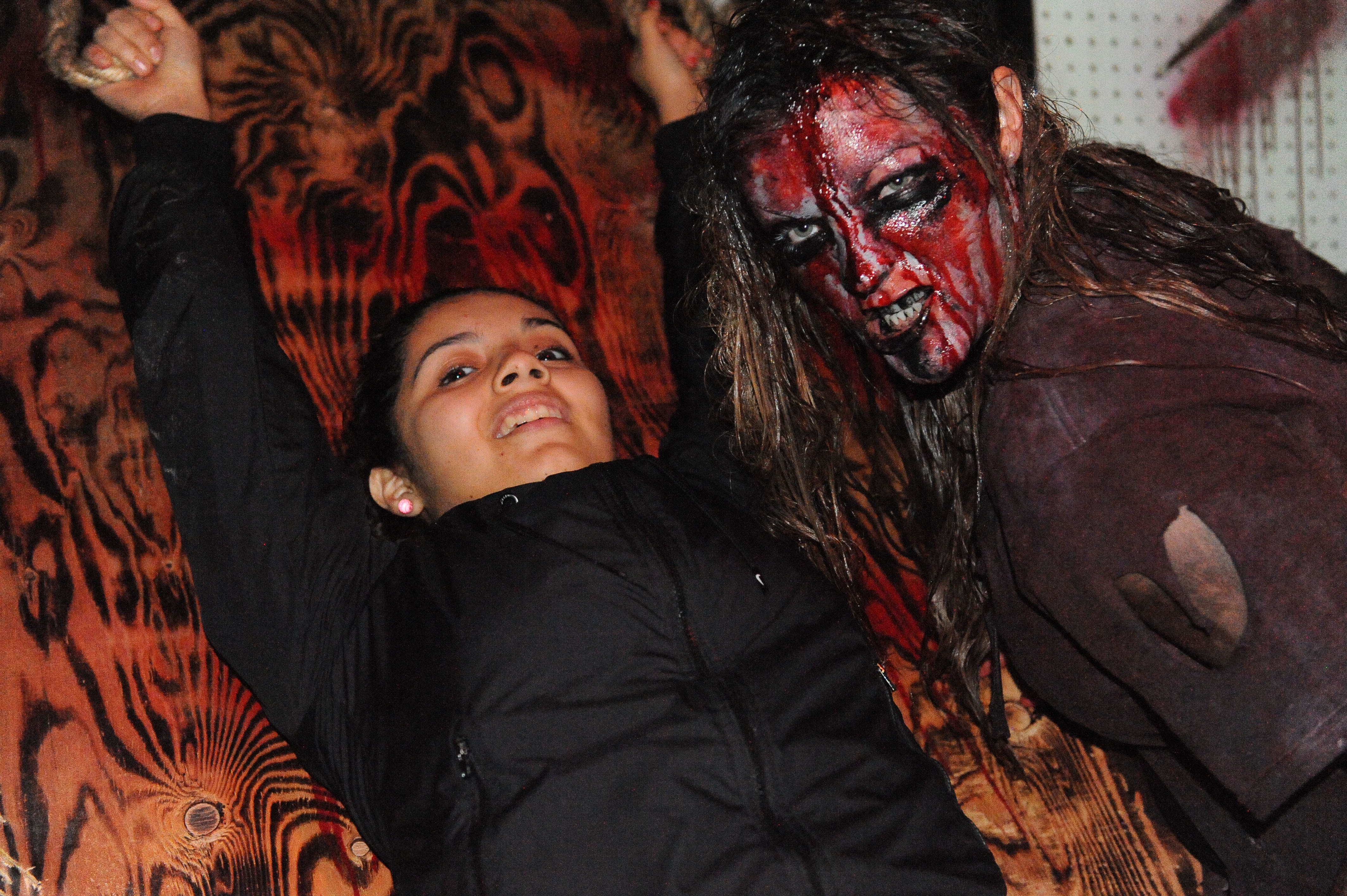 A zombie-costumed performer looms beside a smiling visitor at a haunted house.