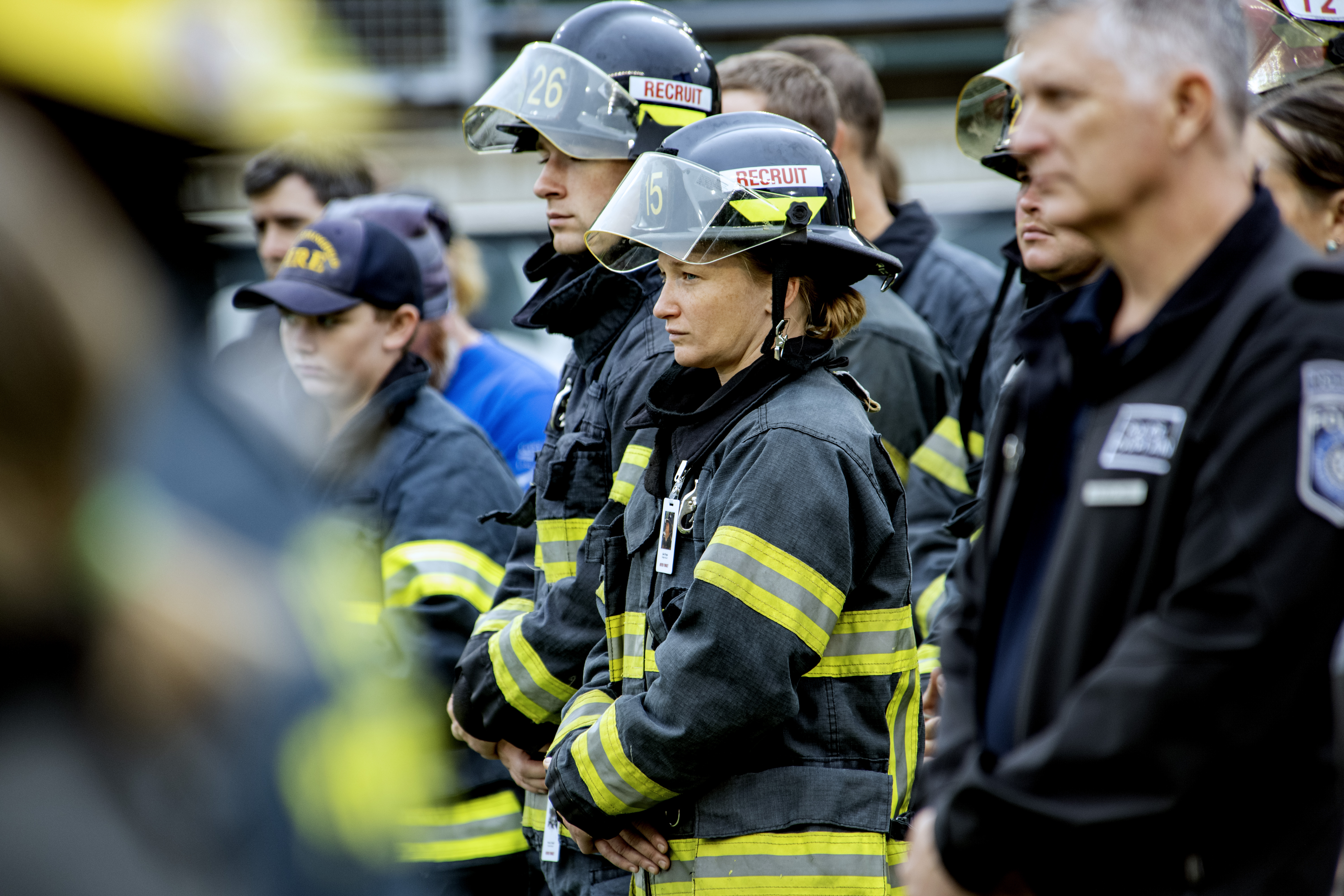 Firefighters in turnout gear and helmets labeled 'Recruit' stand in solemn formation.
