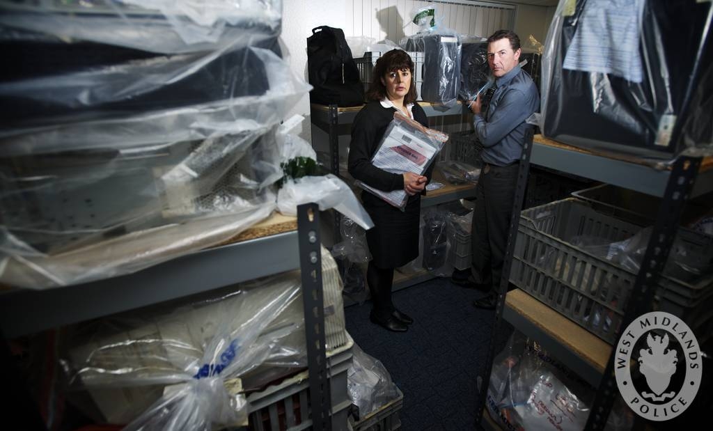 Two officers stand among shelves of seized computers and mobile phones in a police evidence room.