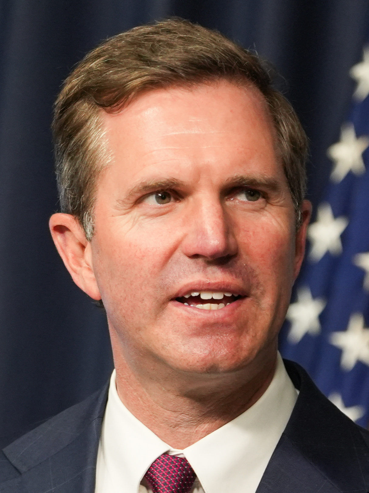 Man in dark suit and burgundy tie speaking at a government podium with an American flag behind him.