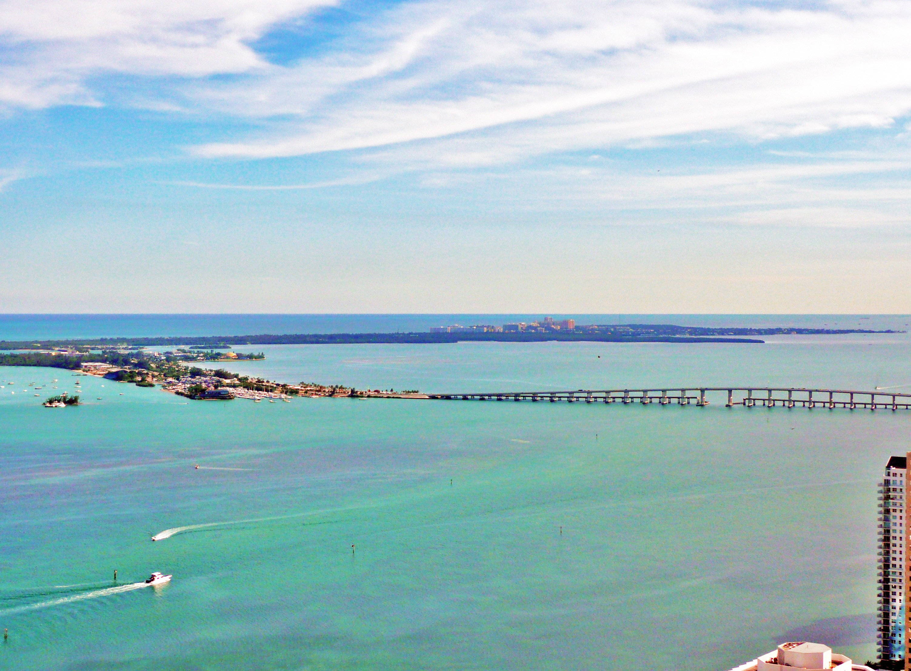 Aerial view of a long causeway bridge over turquoise water connecting an island to a city.