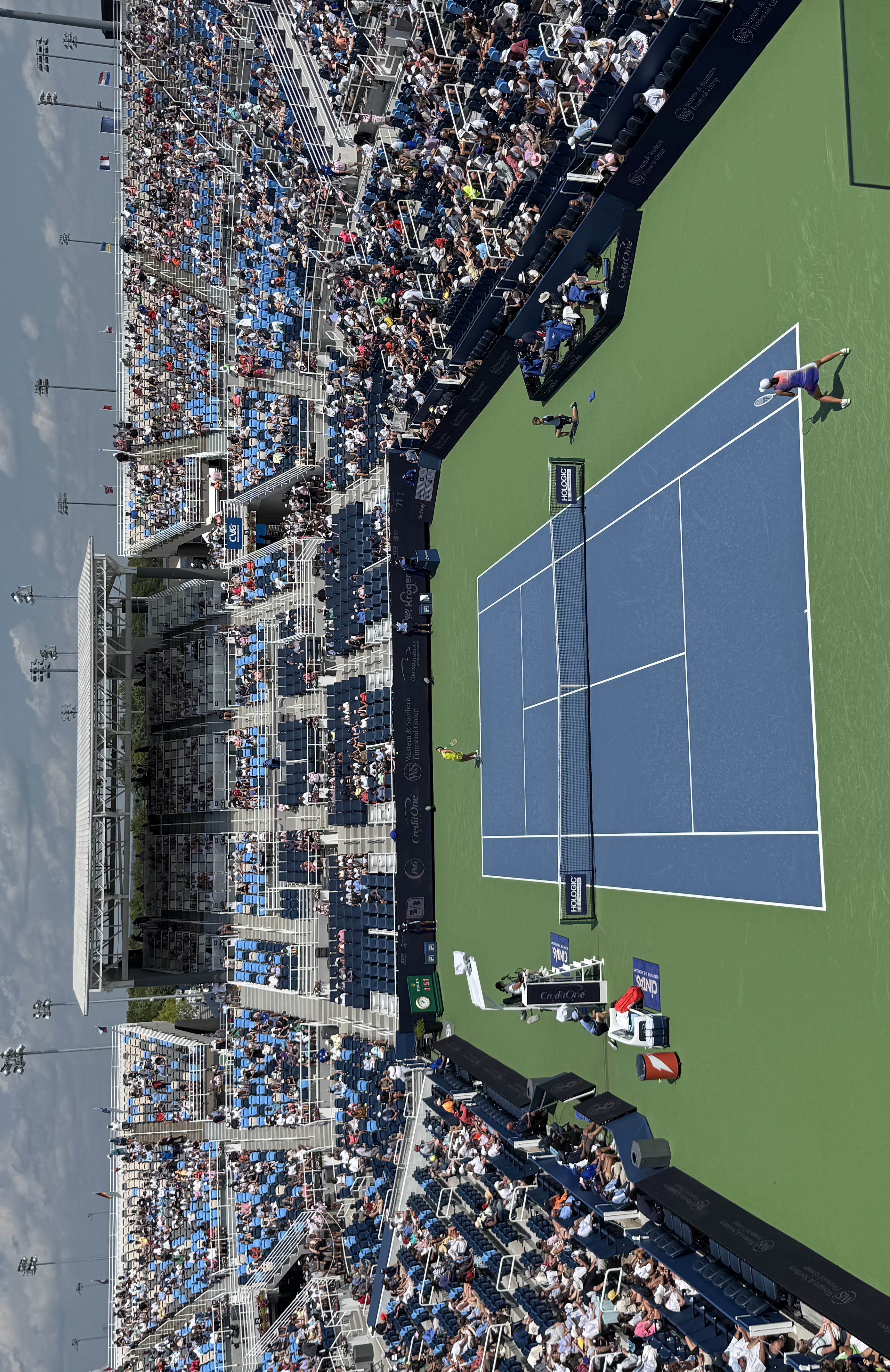Elevated view of two tennis players on a blue hard court surrounded by spectators.