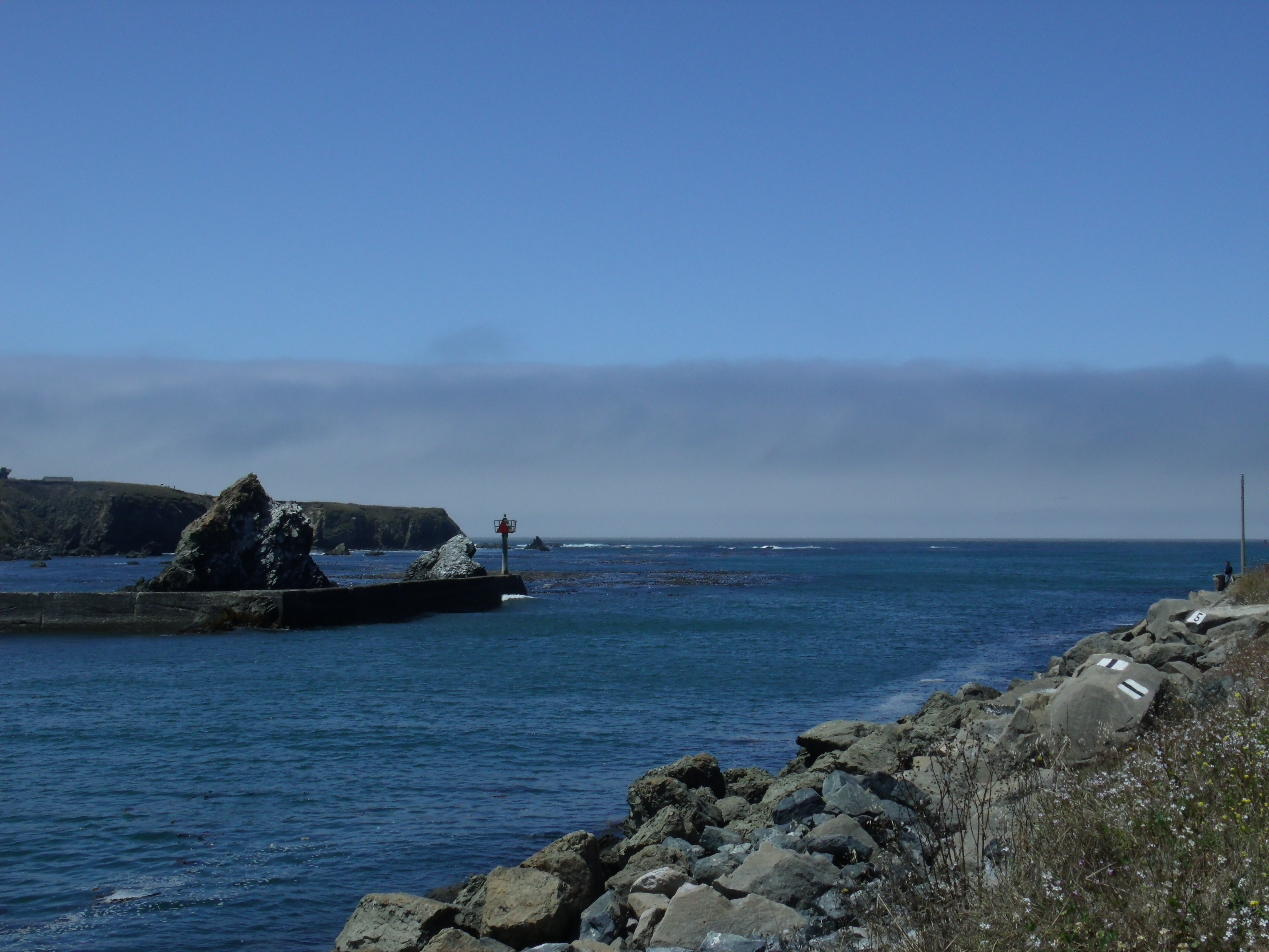 Rocky harbor entrance with calm blue water, a jetty with a red marker, and sea stacks in the distance.