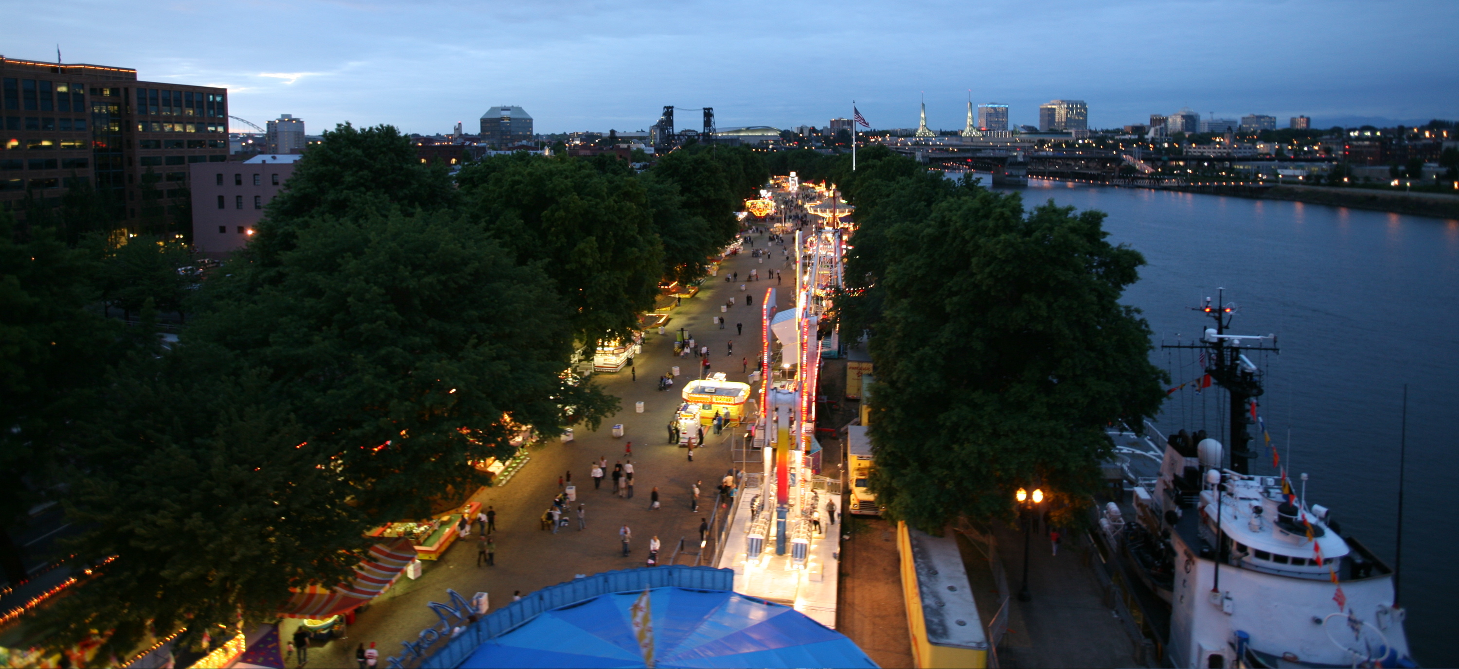 Aerial dusk view of a festival with vendor lights along a riverside promenade.