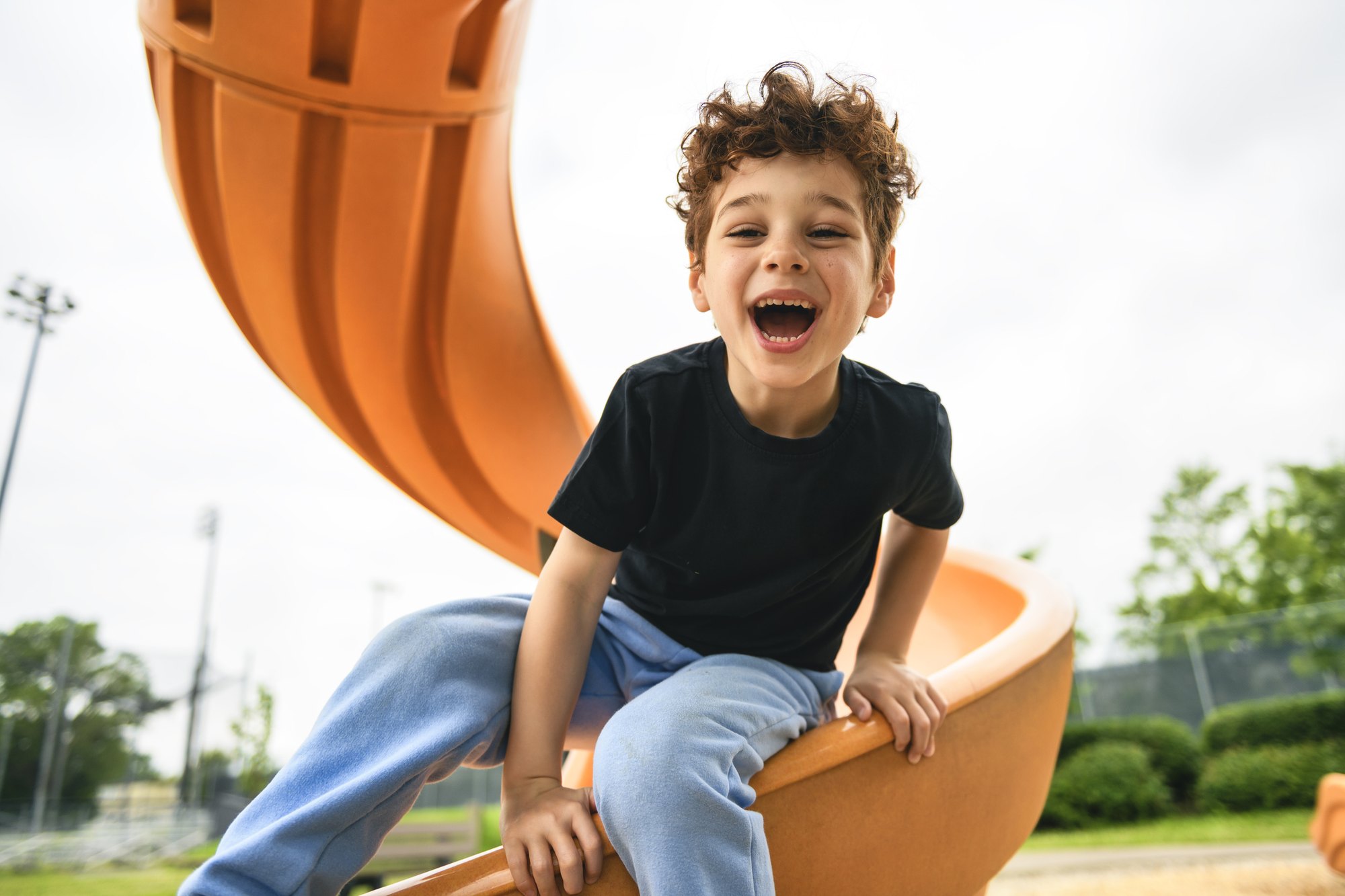 A laughing boy in a black shirt sits on orange playground equipment outdoors.