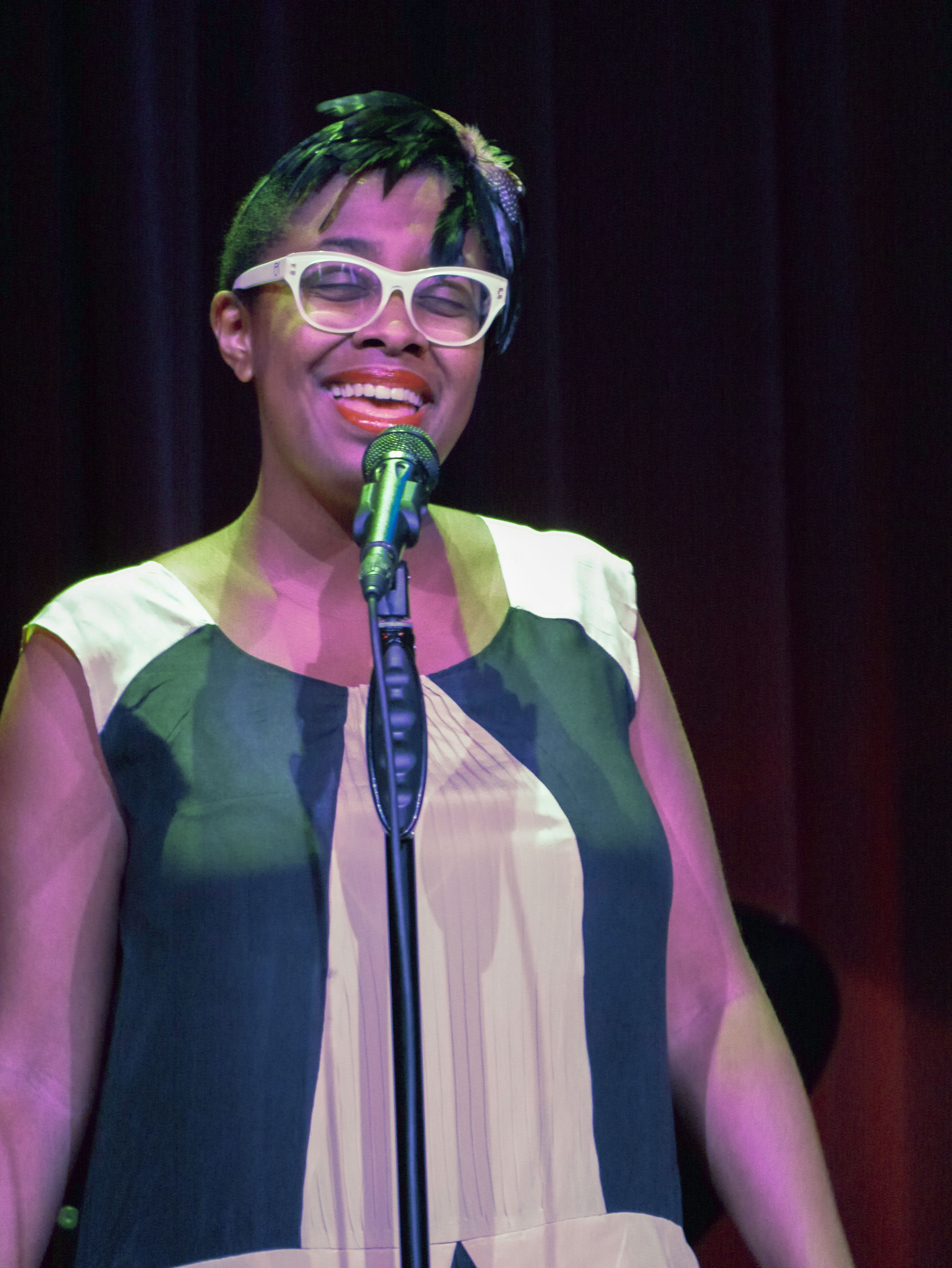 Woman with white-framed glasses singing into a green microphone on a stand against a dark curtain backdrop.