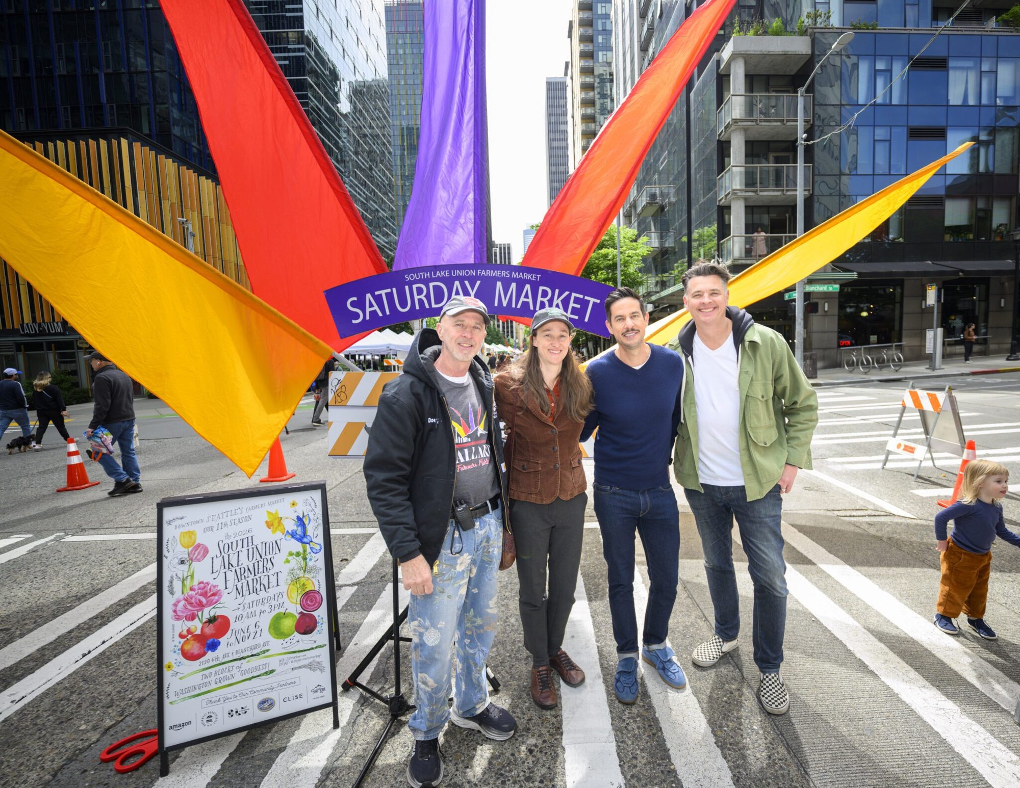 Four adults pose in front of a Saturday Market sign at an outdoor urban farmers market.