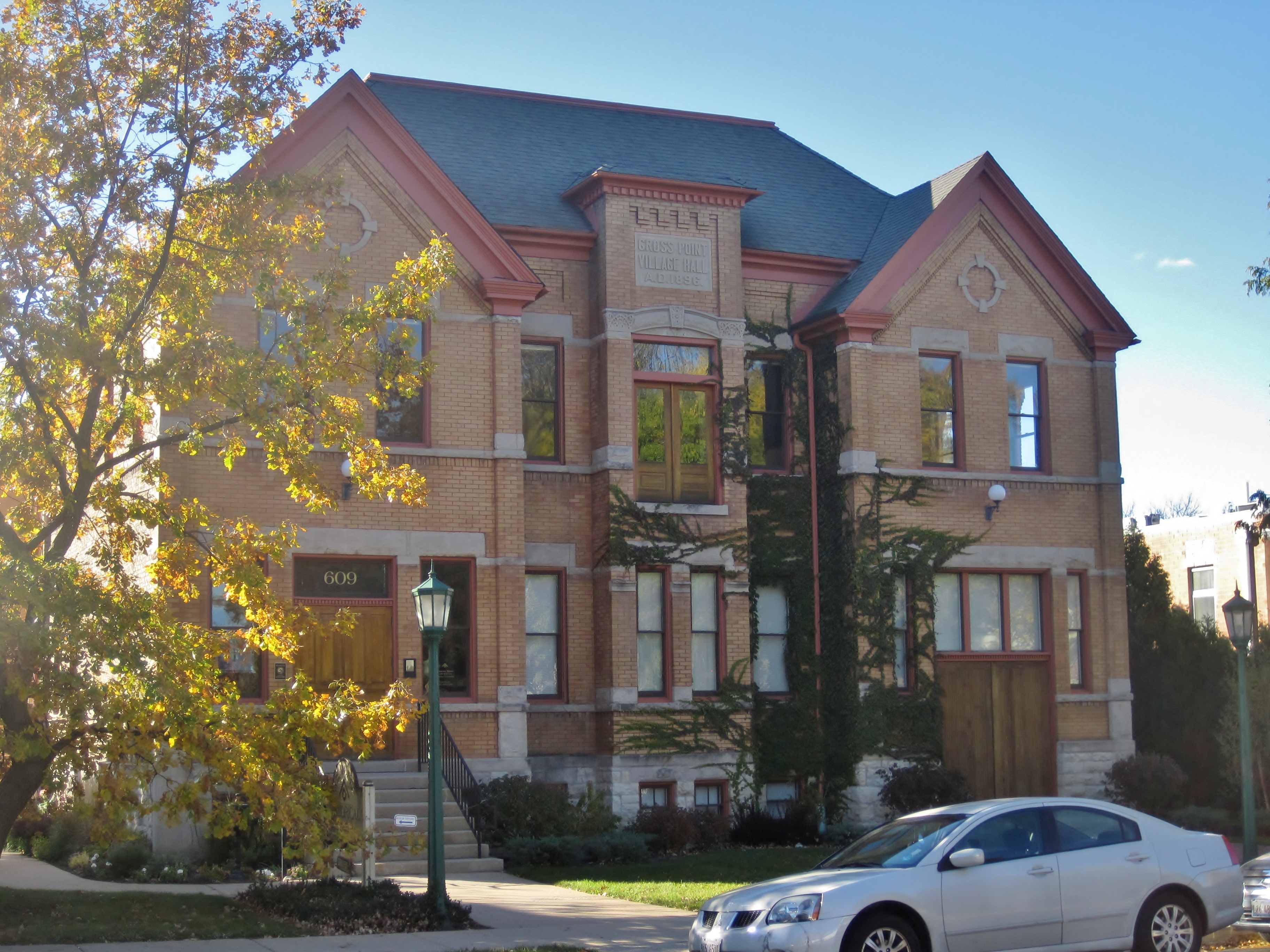 Three-story Victorian brick building with ornate details, autumn tree, and green lamppost on a village street.
