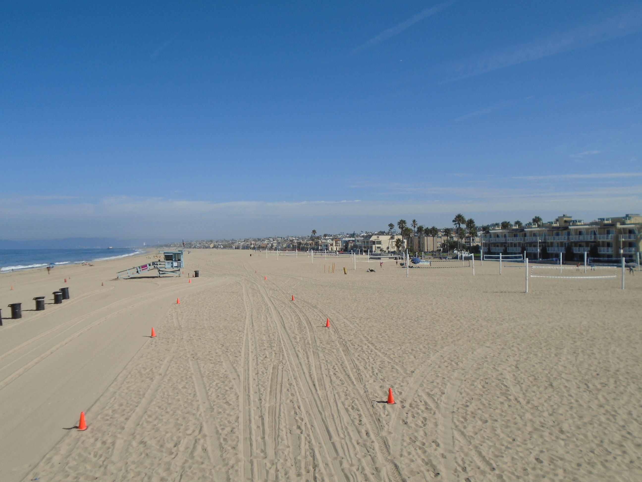 Sandy beach with volleyball nets, a lifeguard tower, and beachfront buildings under a clear sky.
